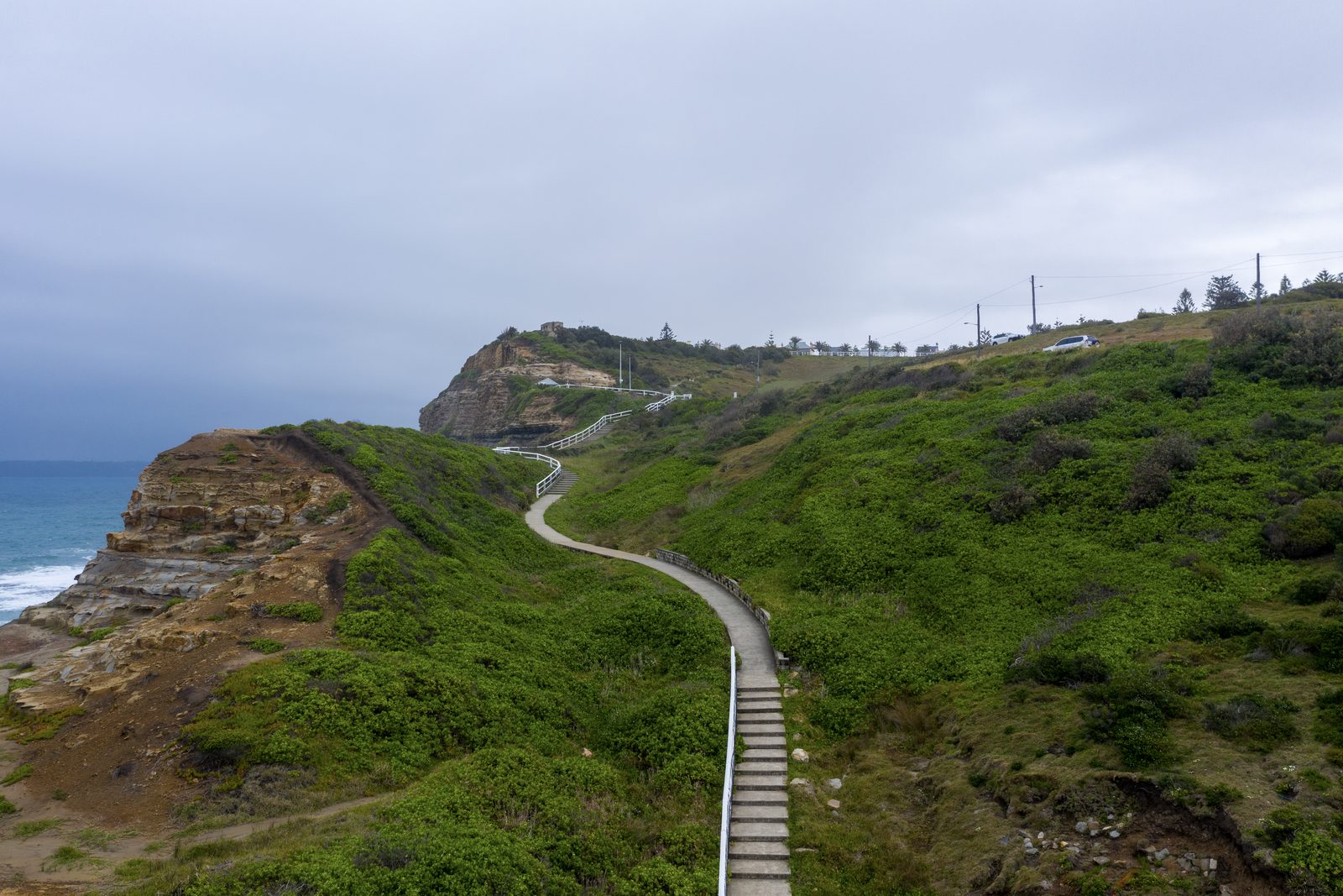 A timber stairway winds up a green coastal headland toward buildings on the ridge, sandstone cliffs and grey sea to the left.