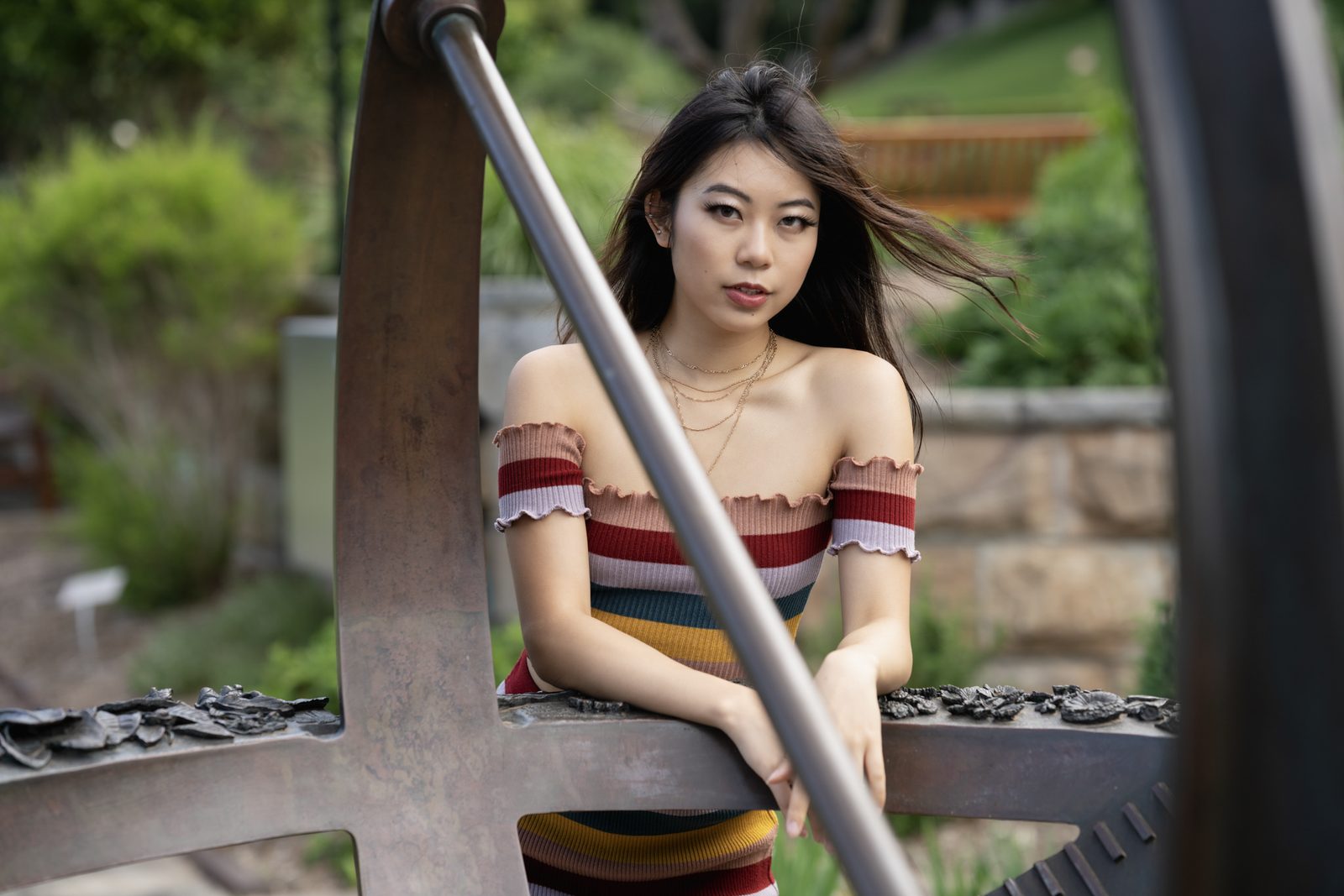 A woman in a striped off-shoulder knit top leans on the spokes of a large rusted iron wheel, a garden soft behind her