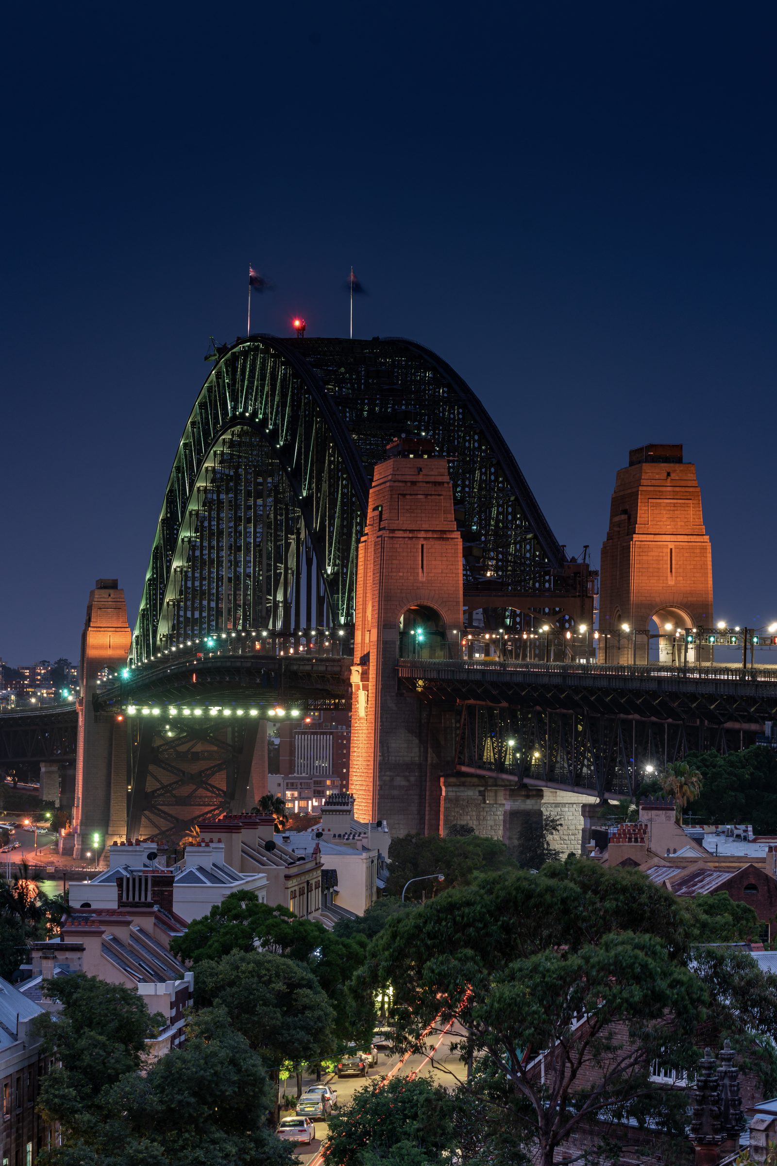 The Sydney Harbour Bridge stands lit against a deep blue evening sky, its sandstone pylons glowing above the terraces below.