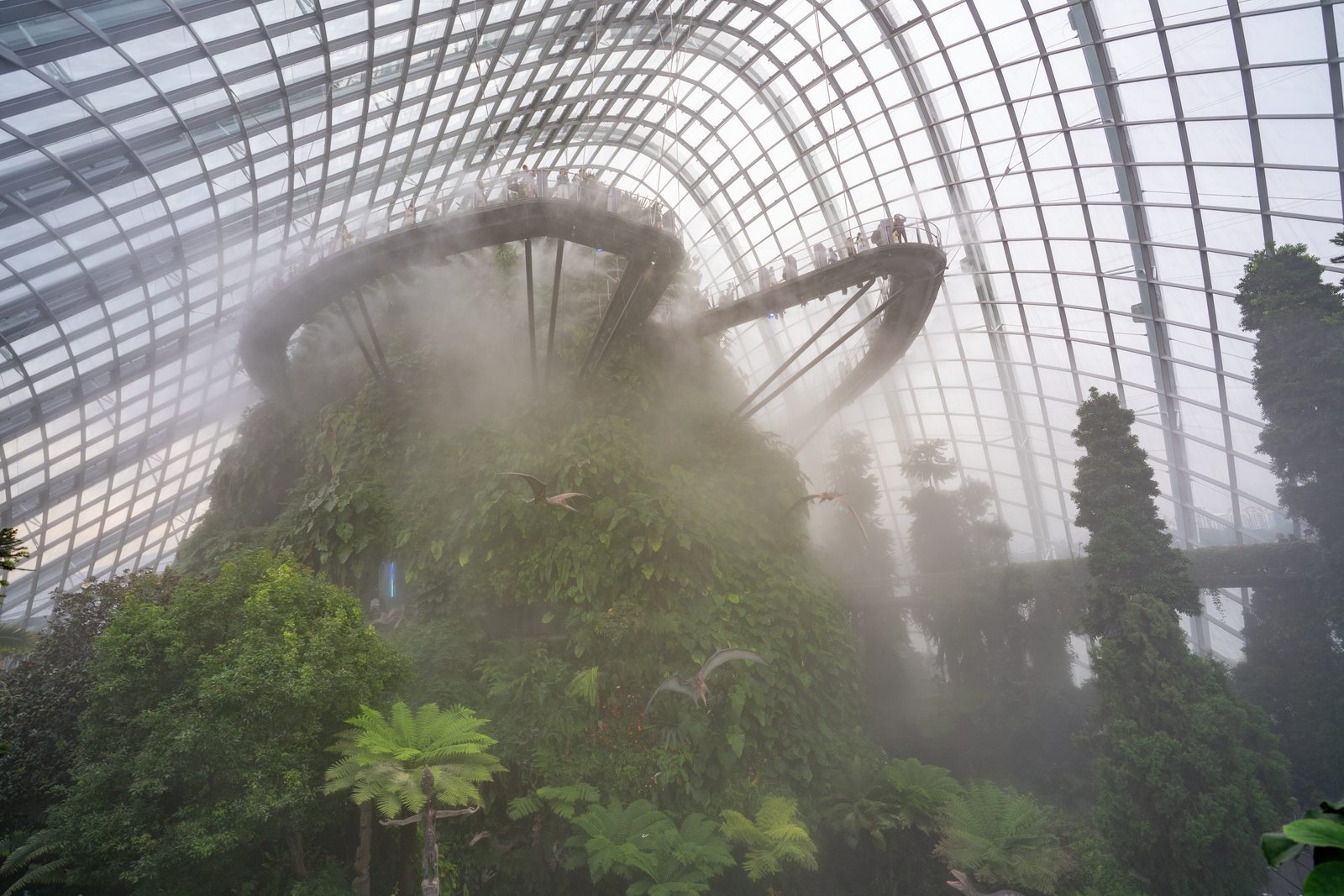 Mist drifts across a planted mountain inside a glass dome, a looping walkway crowded with visitors high above it.