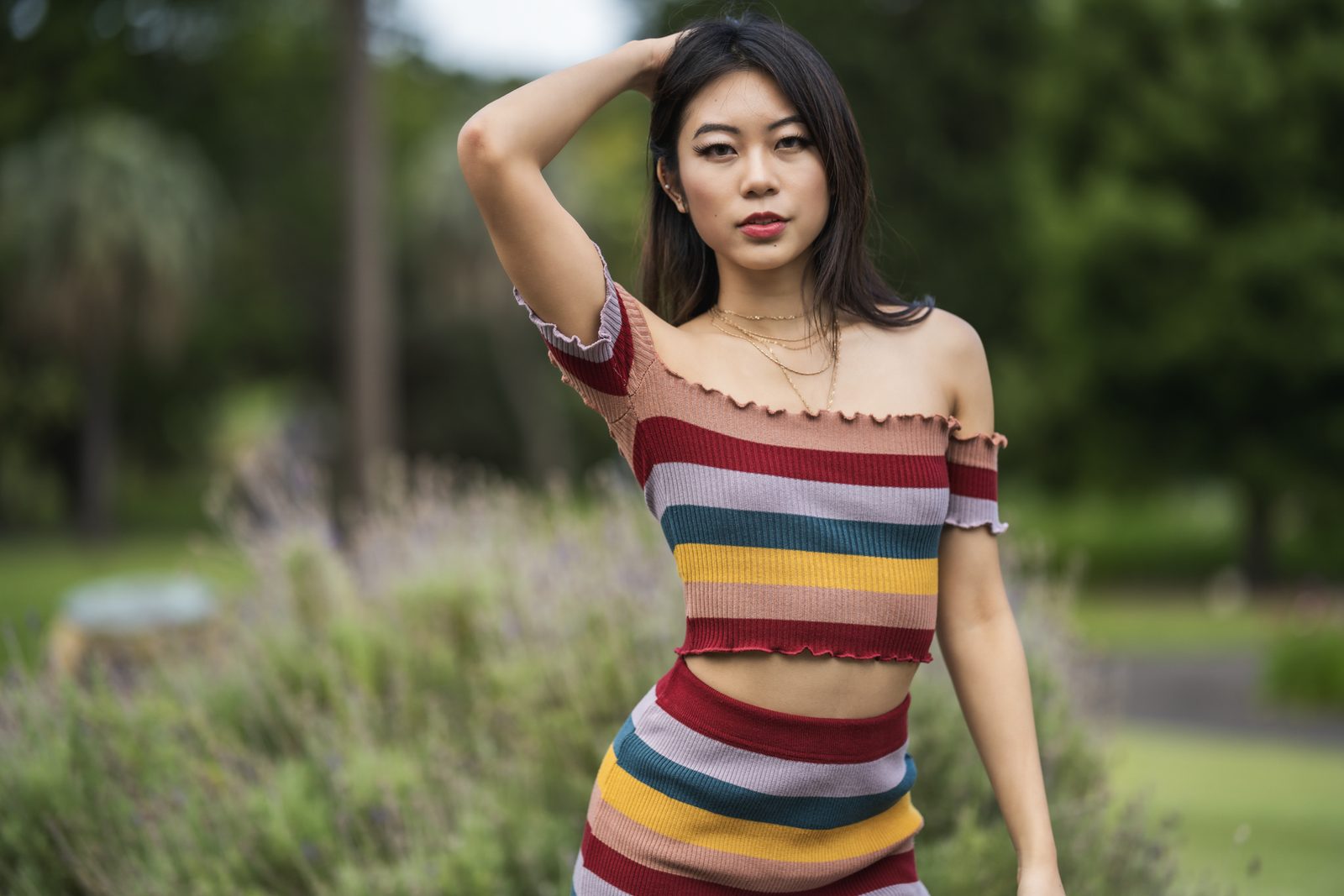 A woman in a striped off-shoulder knit set raises one hand to her hair, lavender bushes blurred behind her