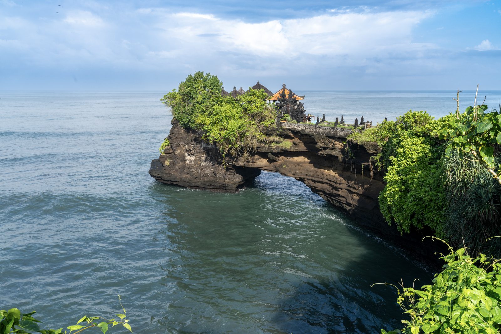 A small temple sits on a rock outcrop pierced by a sea arch, green scrub spilling over the stone above the water.