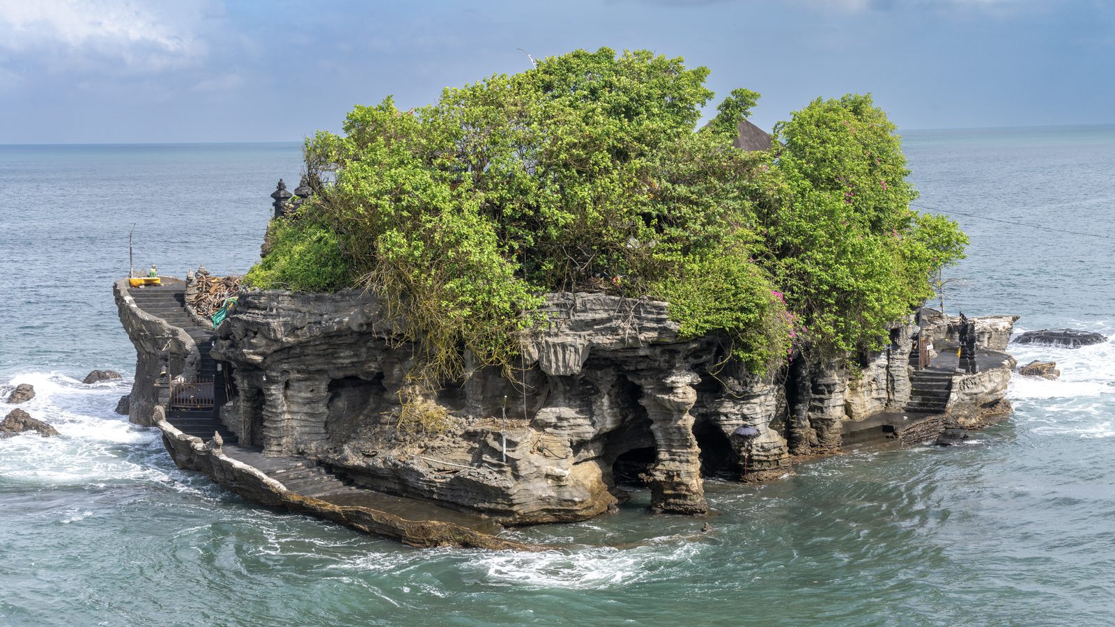 A rock island crowded with trees and shrines rises straight out of the sea, waves breaking around its undercut base.