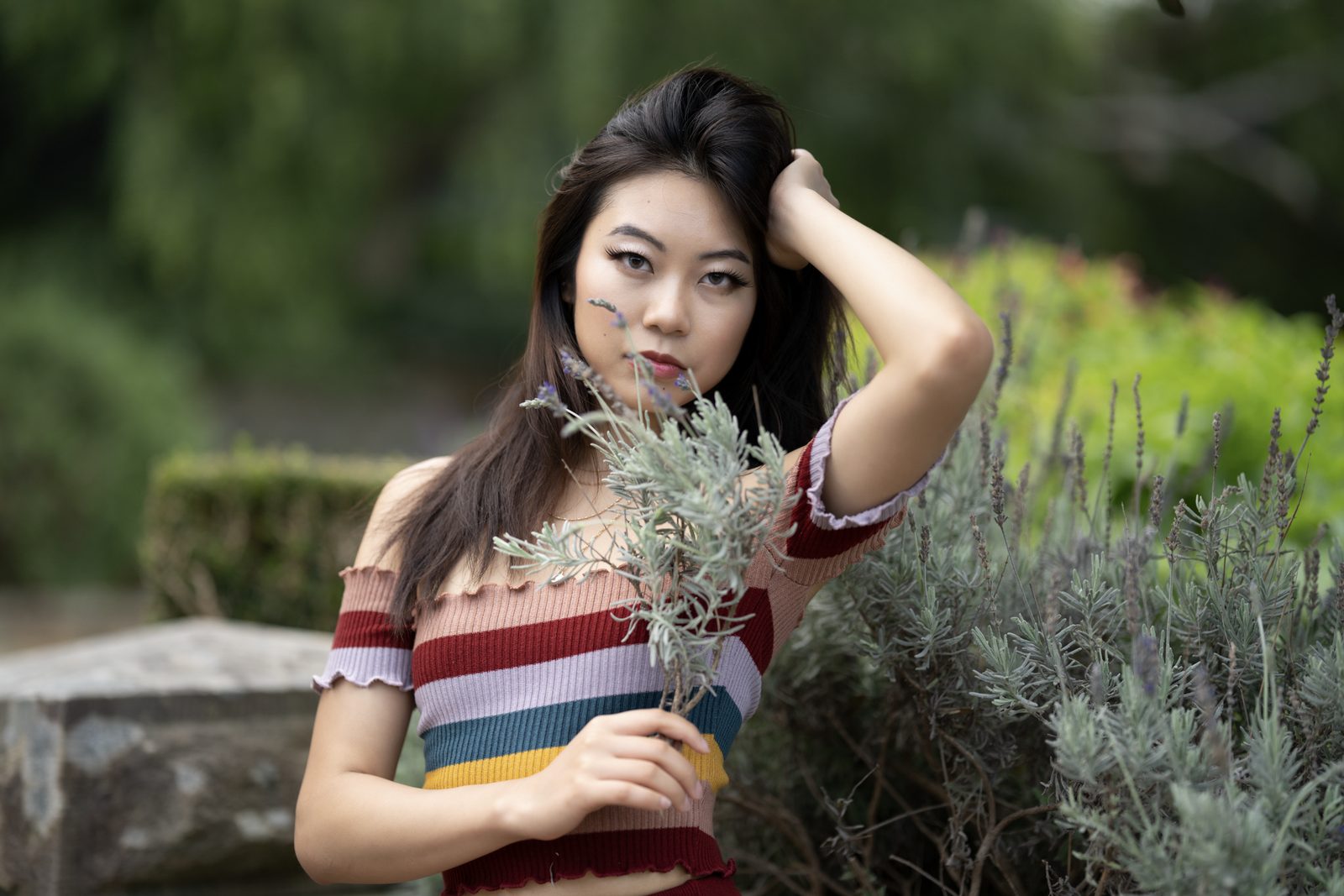 A woman in a striped off-shoulder top holds a sprig of lavender near her face, one hand lifted to her hair