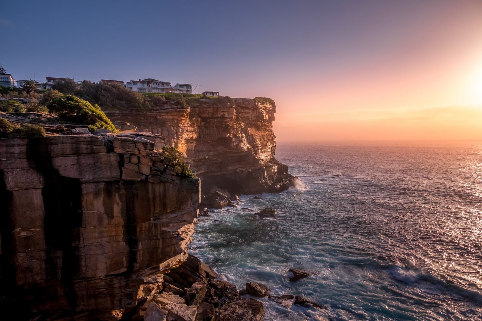 Layered sandstone cliffs drop to a churning sea at sunrise, with houses perched along the clifftop above.