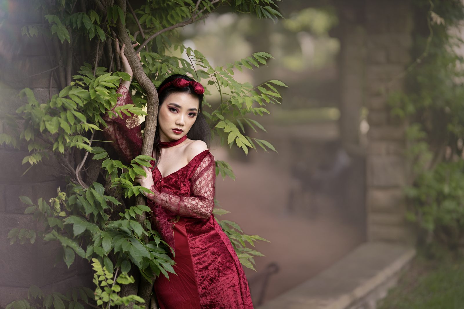 A woman in a red velvet and lace dress with a bow headband leans against a vine-covered wall, green leaves across her shoulder