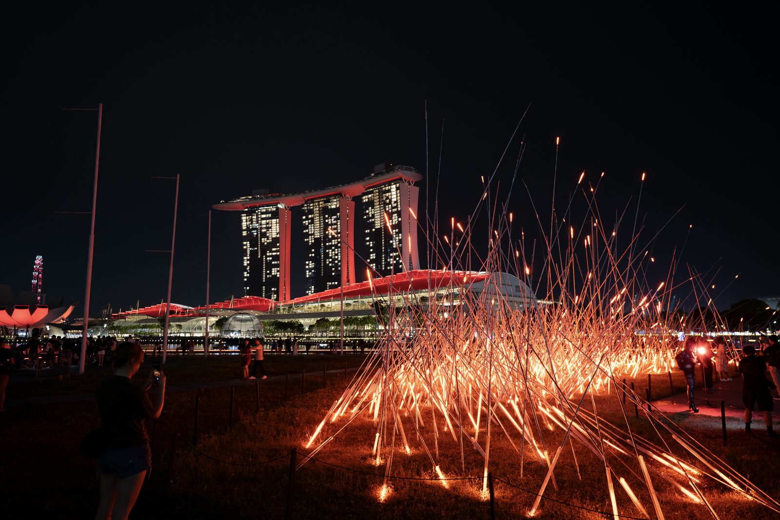 Sparks fountain from the grass in the foreground while a three-towered hotel and lit pavilions stand across the dark bay.