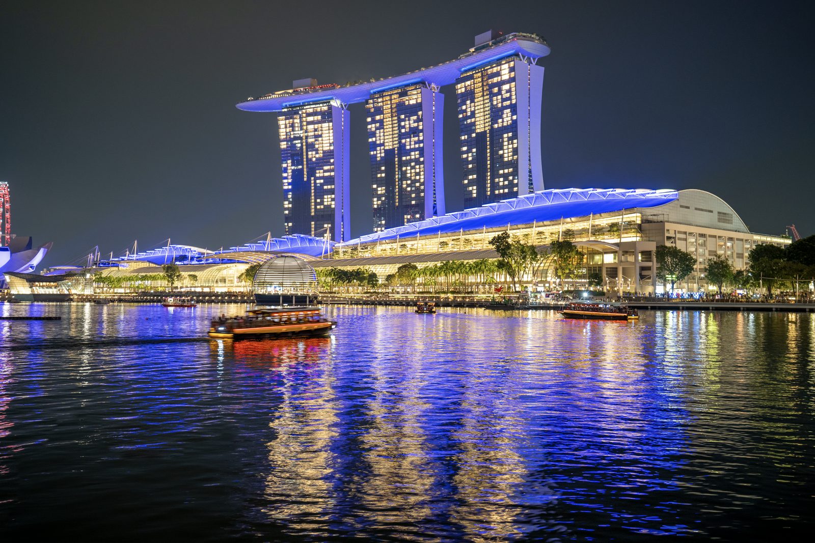 A three-towered hotel and its lit waterfront pavilions stand across still water at night, blue light streaking the reflections.