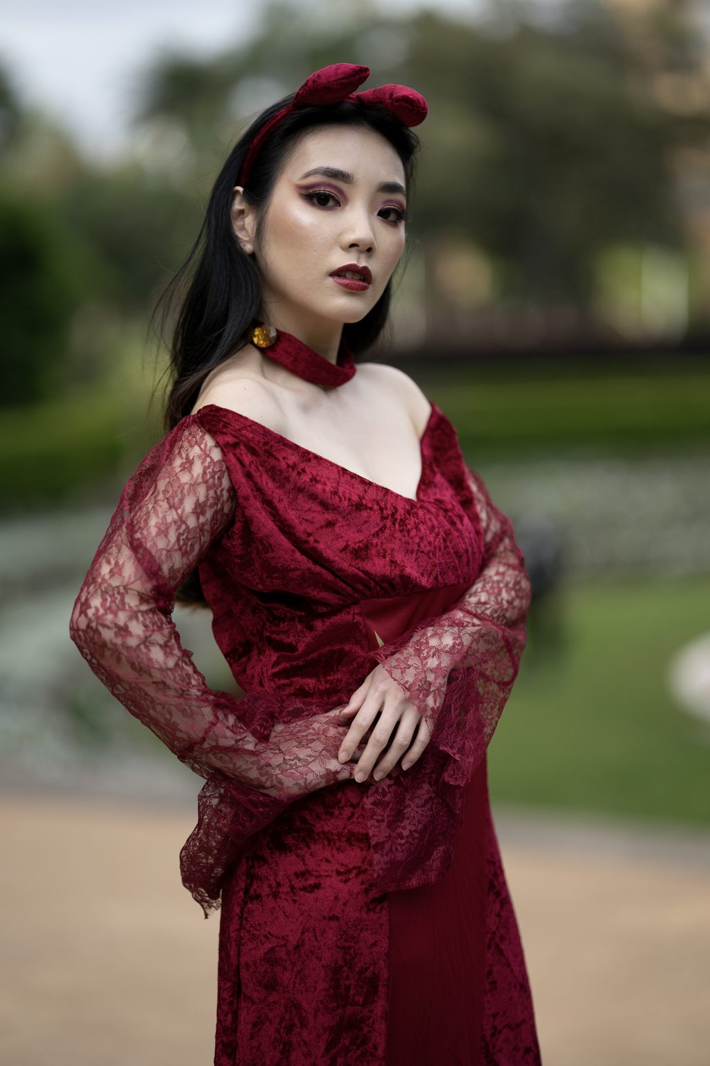A woman in a red velvet and lace dress with a bow headband stands with one hand at her hip, a green park blurred behind