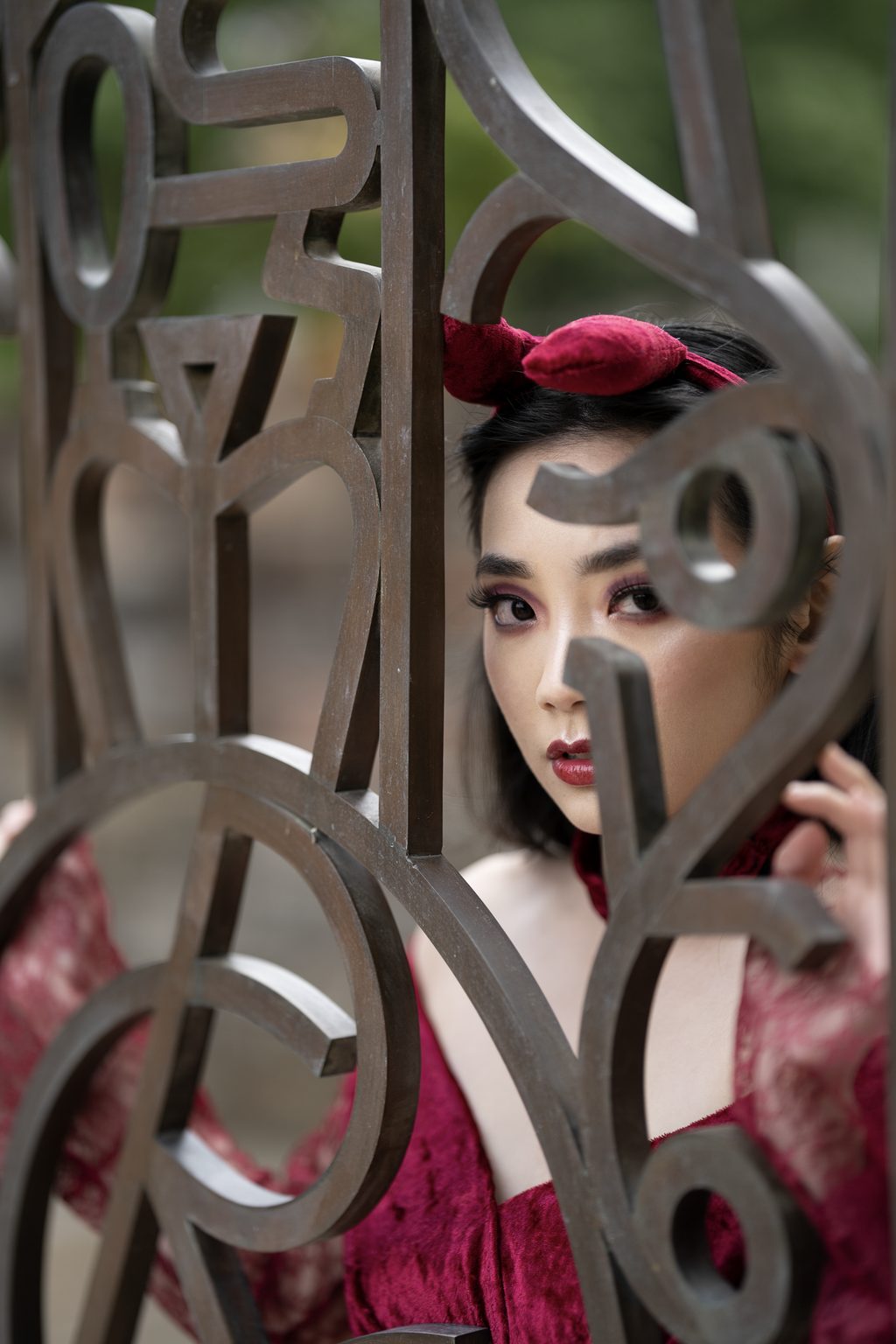 A woman in red looks out between the curls of a rusted decorative metal screen, one hand raised against it