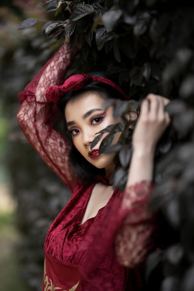 A woman in a red lace dress raises one arm into dark foliage, her face turned to the camera between the leaves