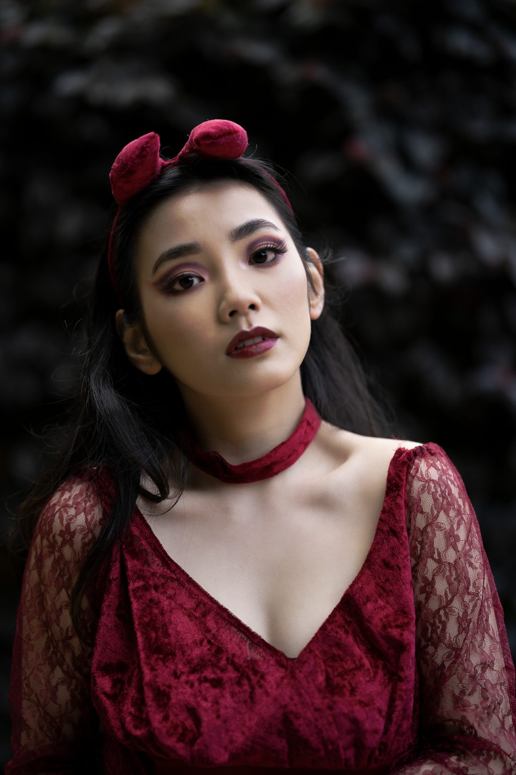 A close portrait of a woman in red velvet with a matching choker and bow headband, dark leaves behind her