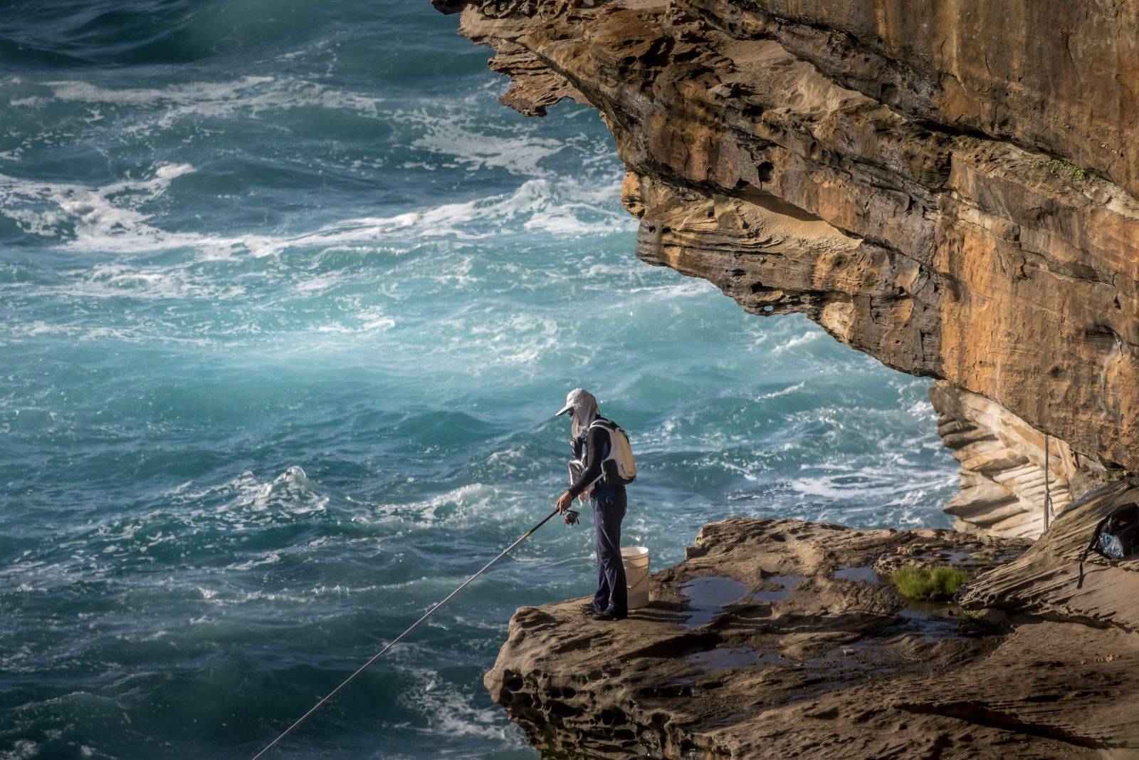 A lone fisherman casts from a sandstone ledge above churning turquoise water, a great cliff overhang above him.