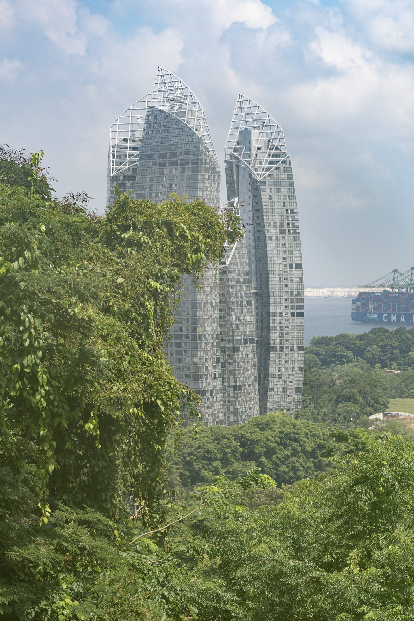 Two curved glass towers rise beyond dense green foliage, a container ship on the hazy water behind them.