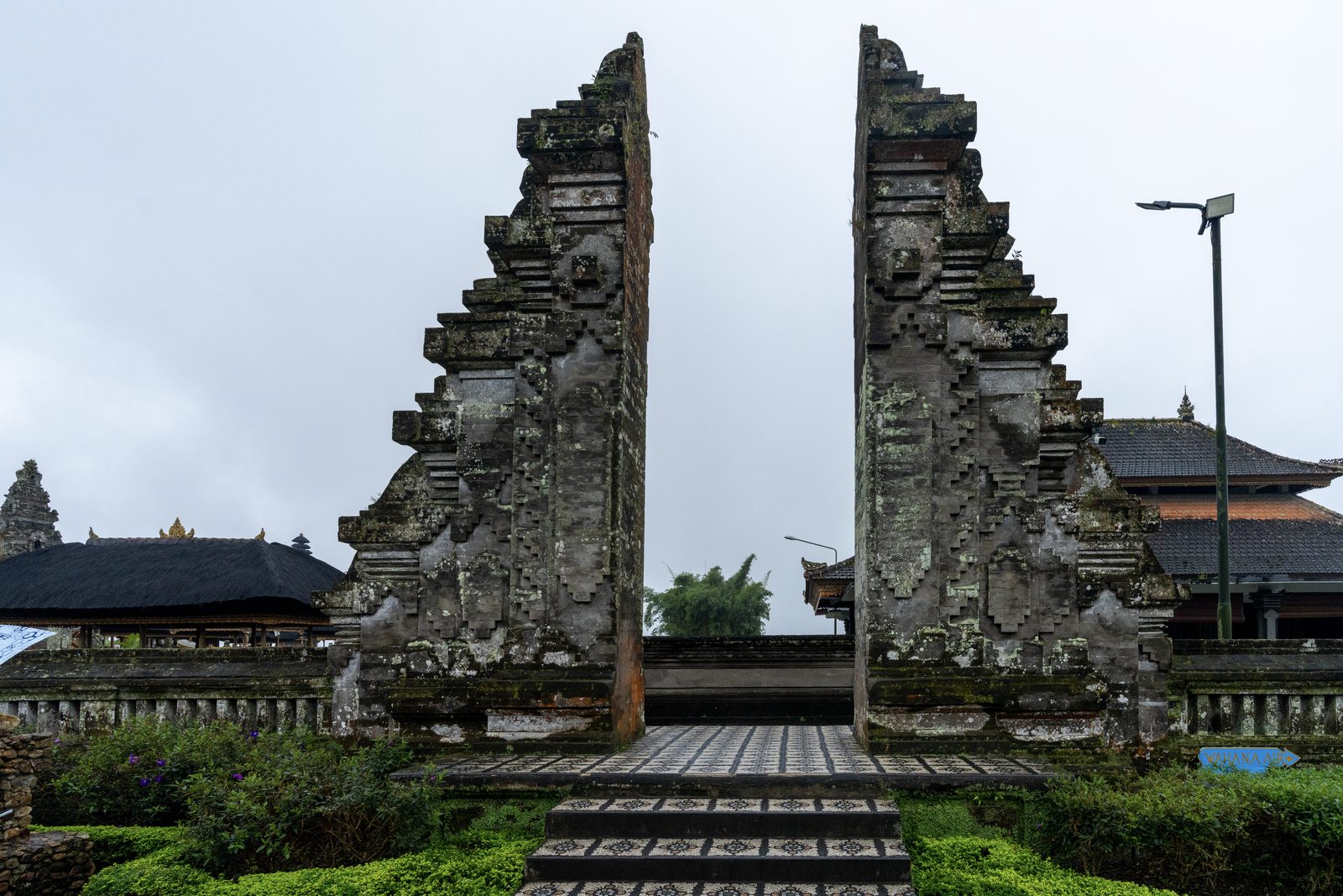 A moss-covered split temple gate stands open onto flat grey sky, thatched shrine roofs to either side.