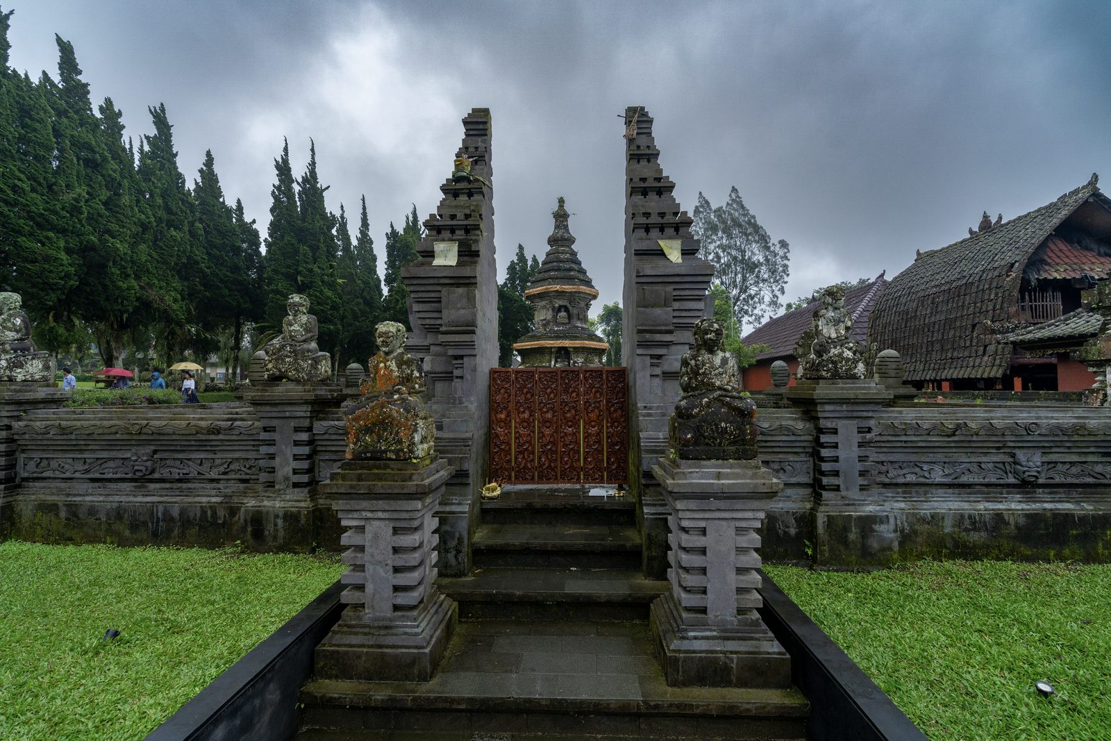 A paved path climbs between carved stone gate posts toward a red and gold door, heavy cloud over the pines behind.