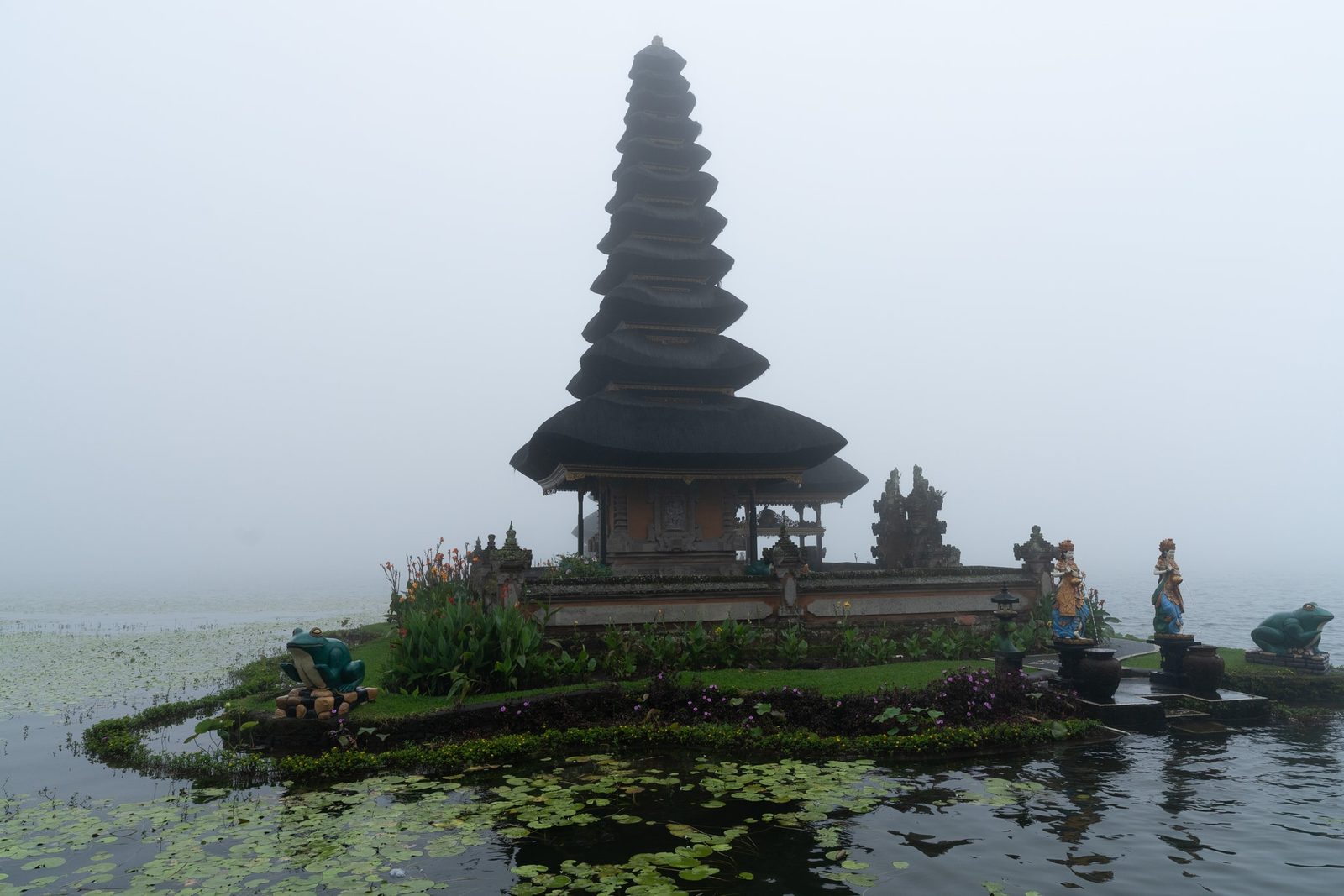 A tiered thatched-roof shrine rises from a lake shrouded in mist, water lilies floating in the foreground.