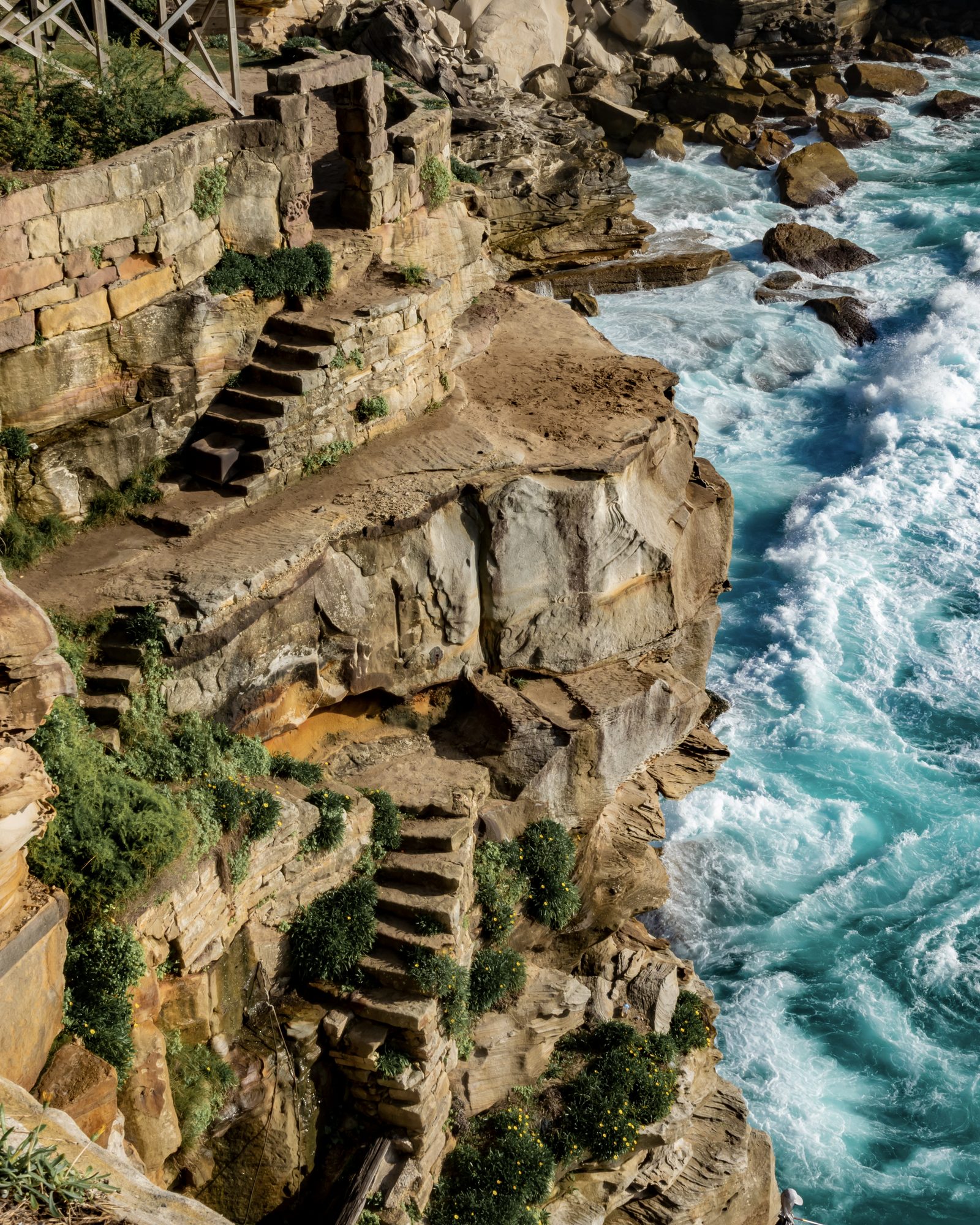 Stone steps cut into a layered sandstone cliff descend toward turquoise water breaking white on the rocks below.