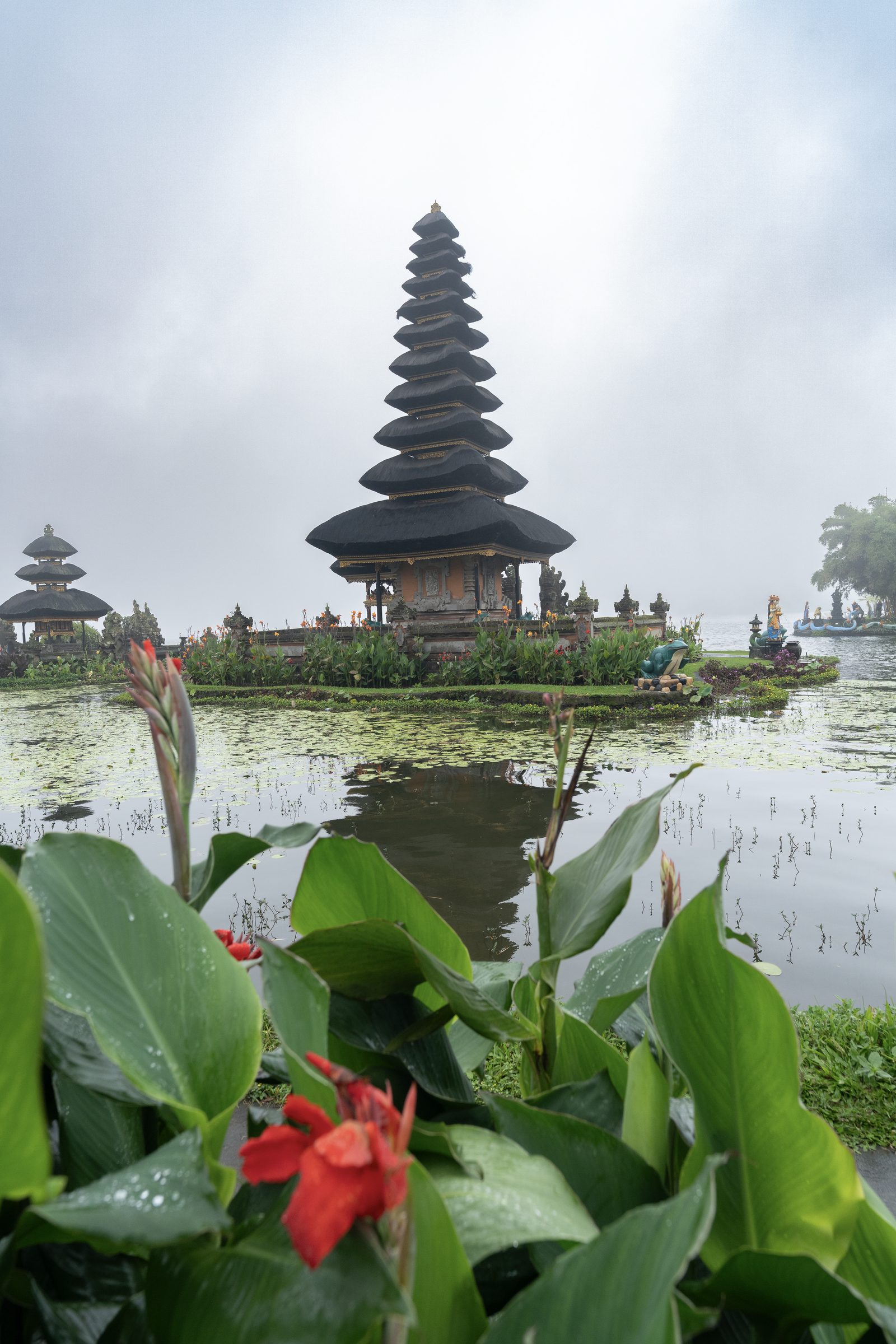 A tiered black-thatched temple sits across a still lake in mist, red canna flowers and wet leaves filling the foreground.