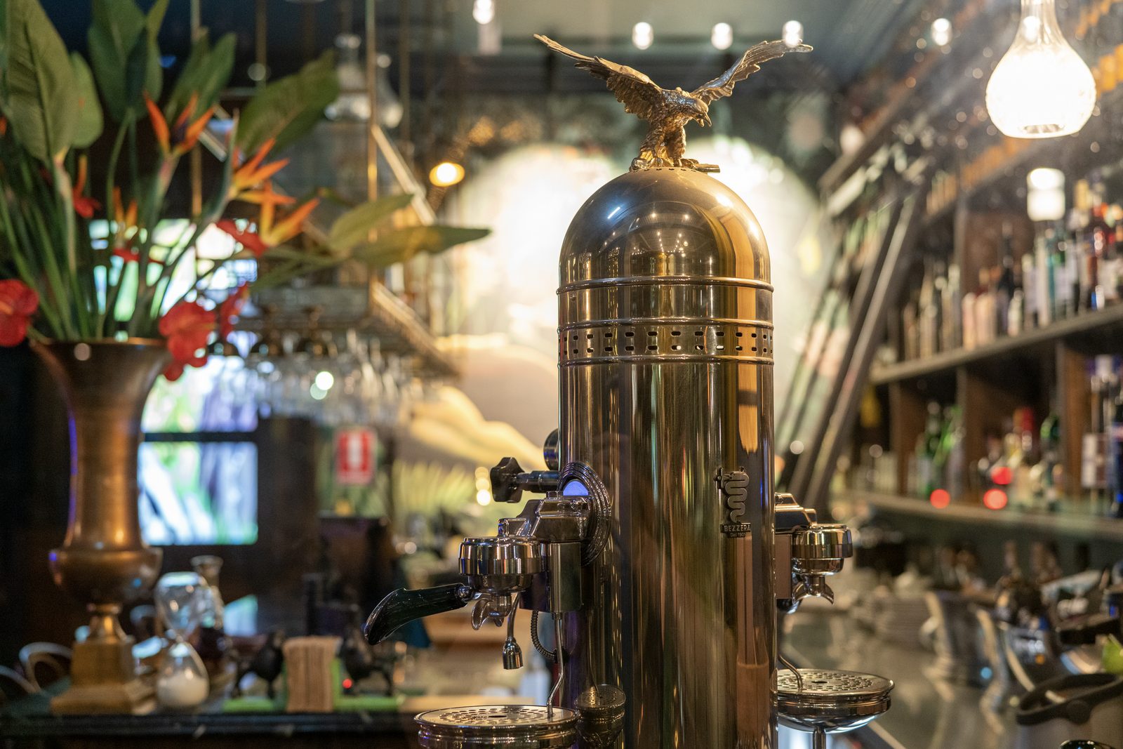 A polished brass beer tower topped with a cast eagle stands on a bar, bottles and warm pendant lights blurred behind it.