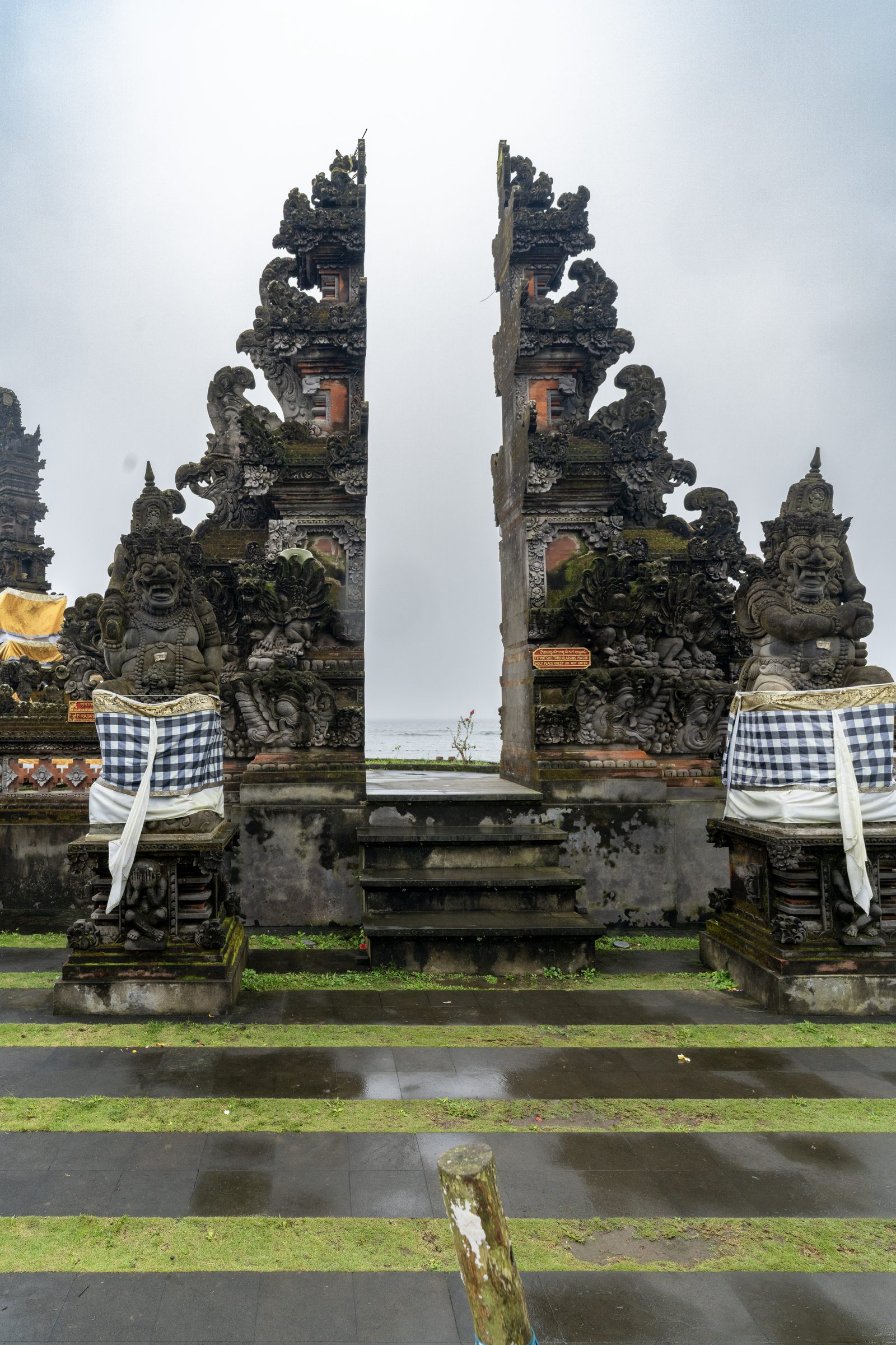 An ornately carved split gate frames flat grey sea, guardian statues to either side wrapped in black and white checked cloth.