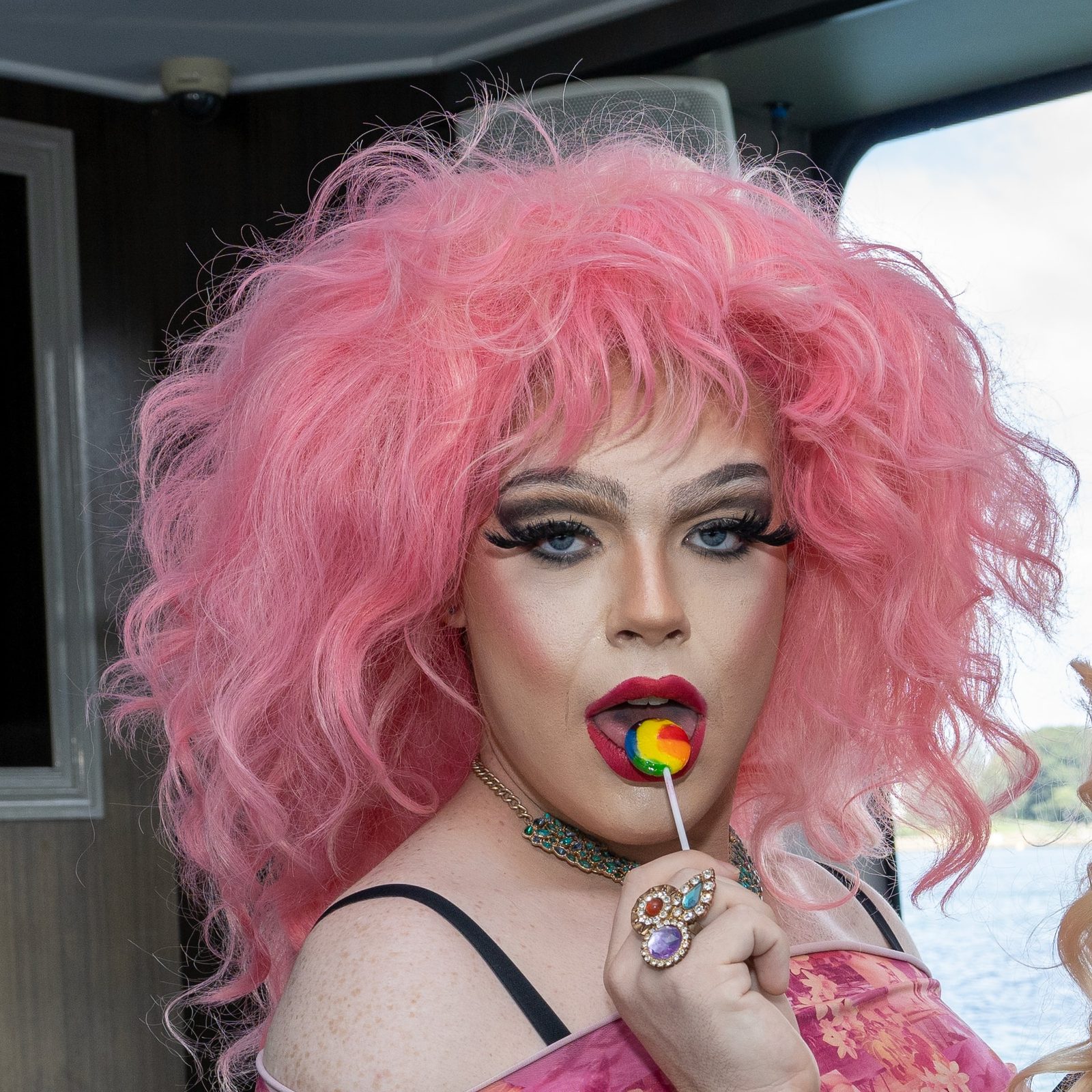 A drag performer with a voluminous pink wig and bold makeup holds a rainbow lollipop to painted lips, jewelled rings on their hand