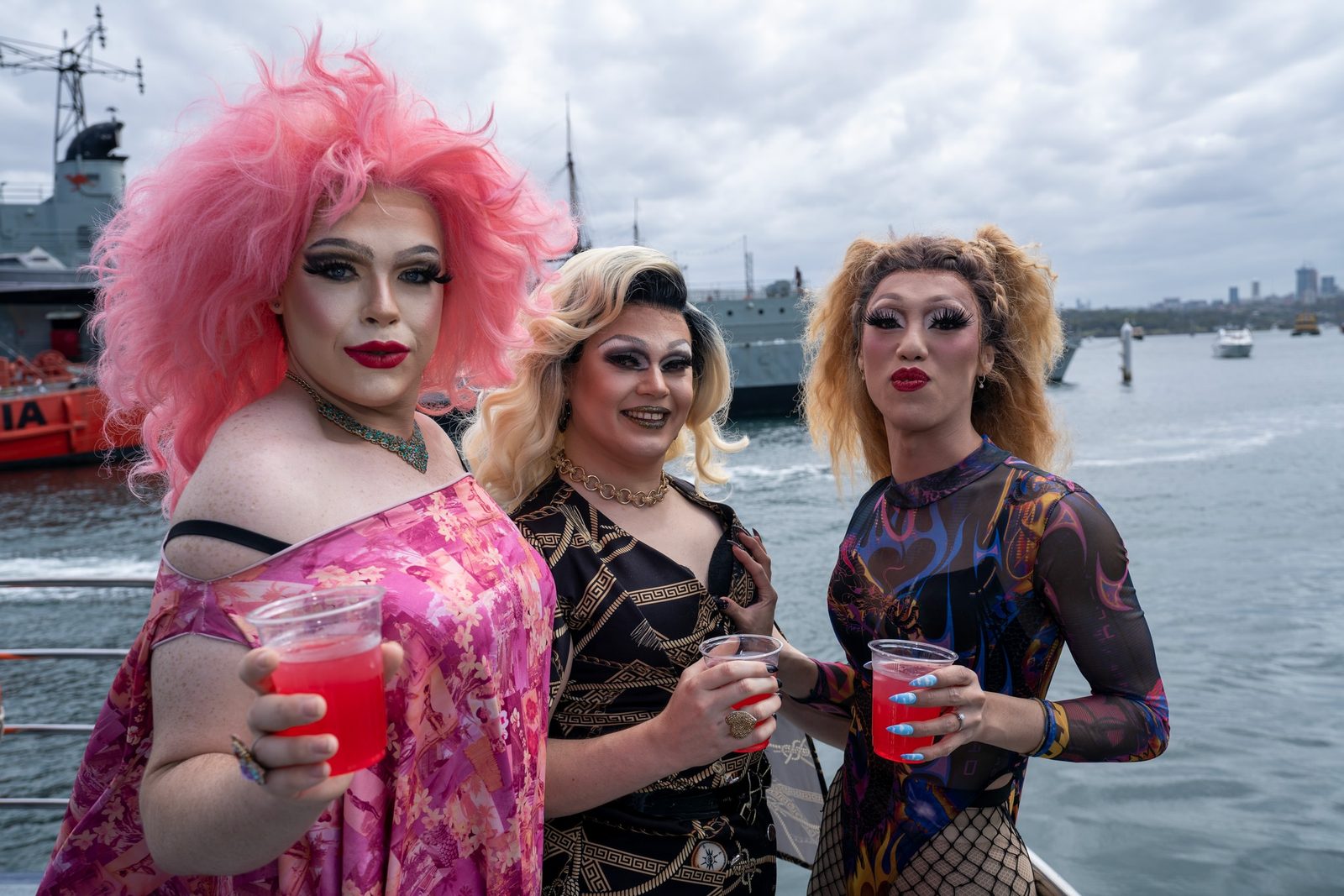 Three drag performers in colourful wigs and makeup stand together on a boat deck, holding drinks, with a grey ship and skyline behind them