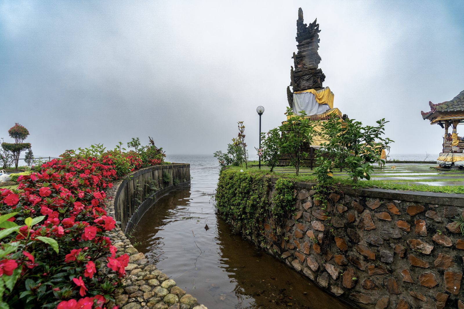 A stone channel of water runs between red flowerbeds toward a gold-robed statue, mist closing off the lake behind.
