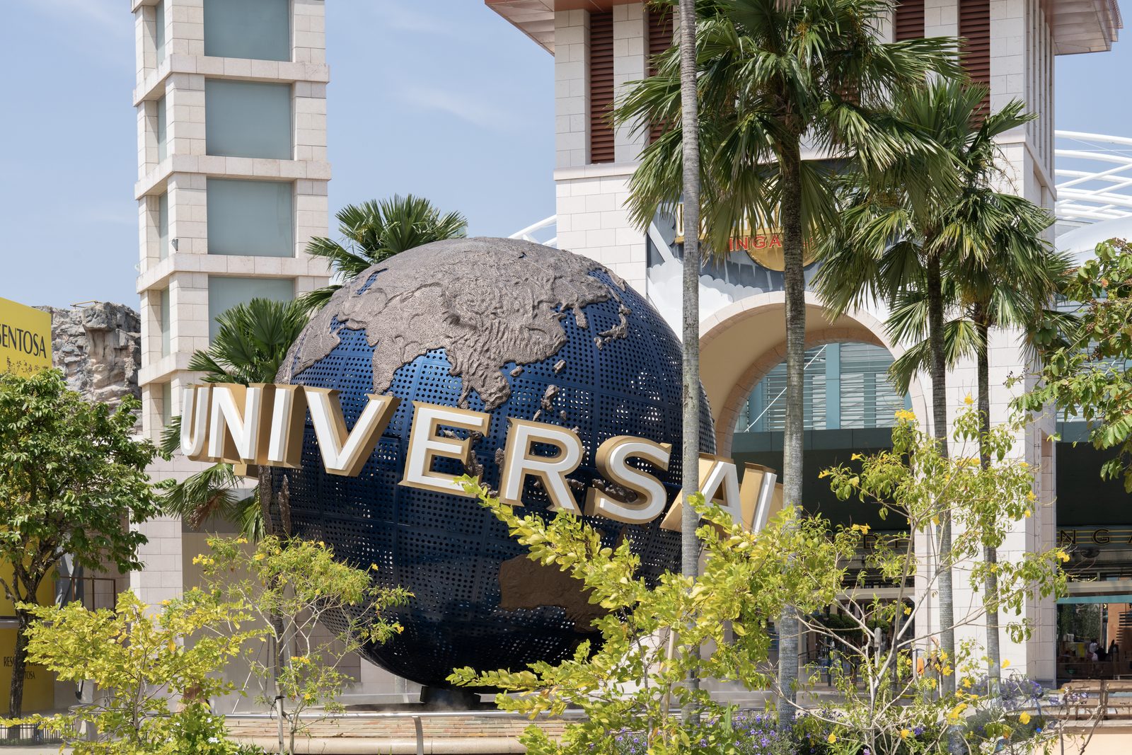 A large metal globe on a plaza spells out a studio name in gold letters, palms and pale buildings around it.