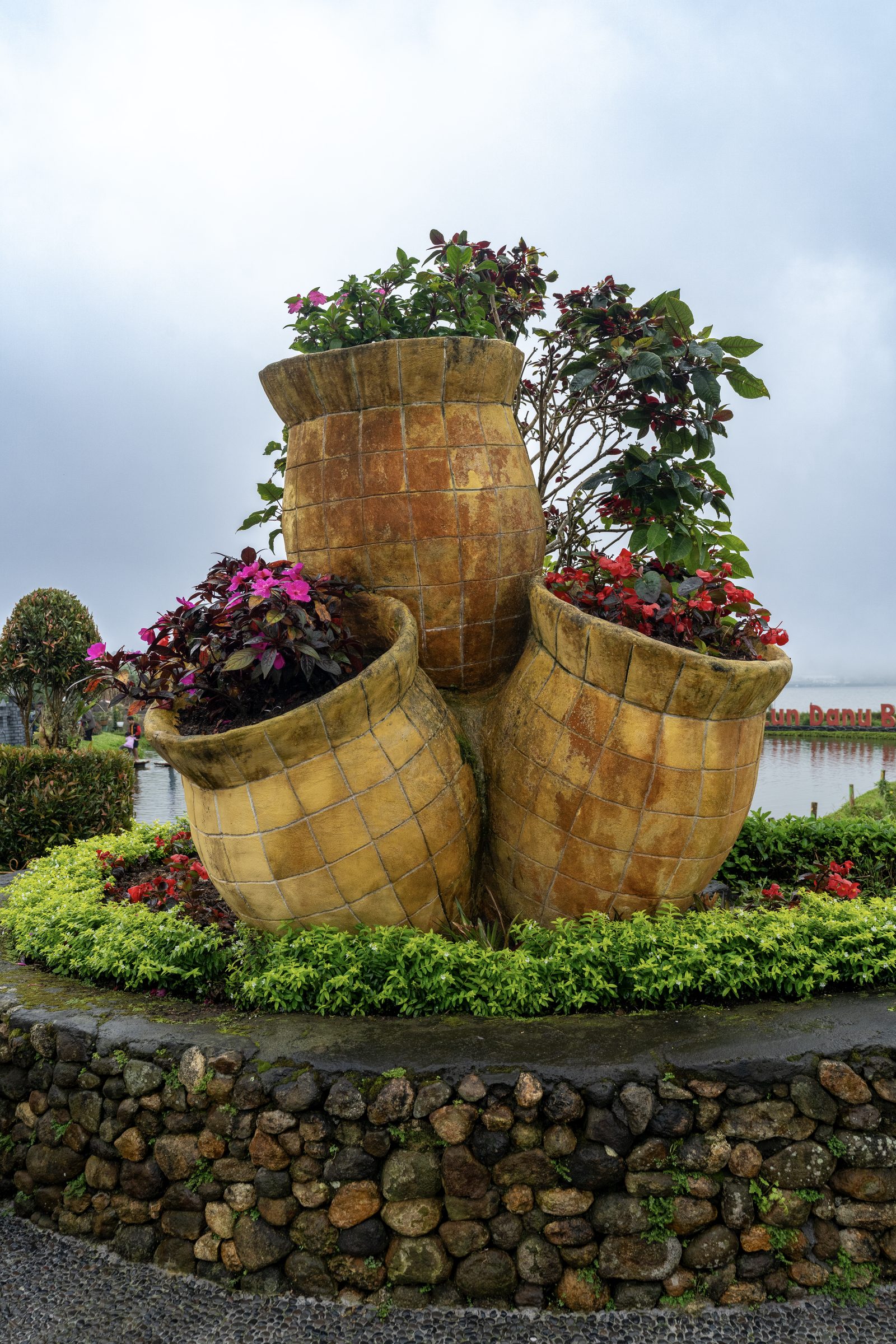 Three oversized tiled pots lean together on a raised bed planted with red and pink flowers, a misty lake behind.