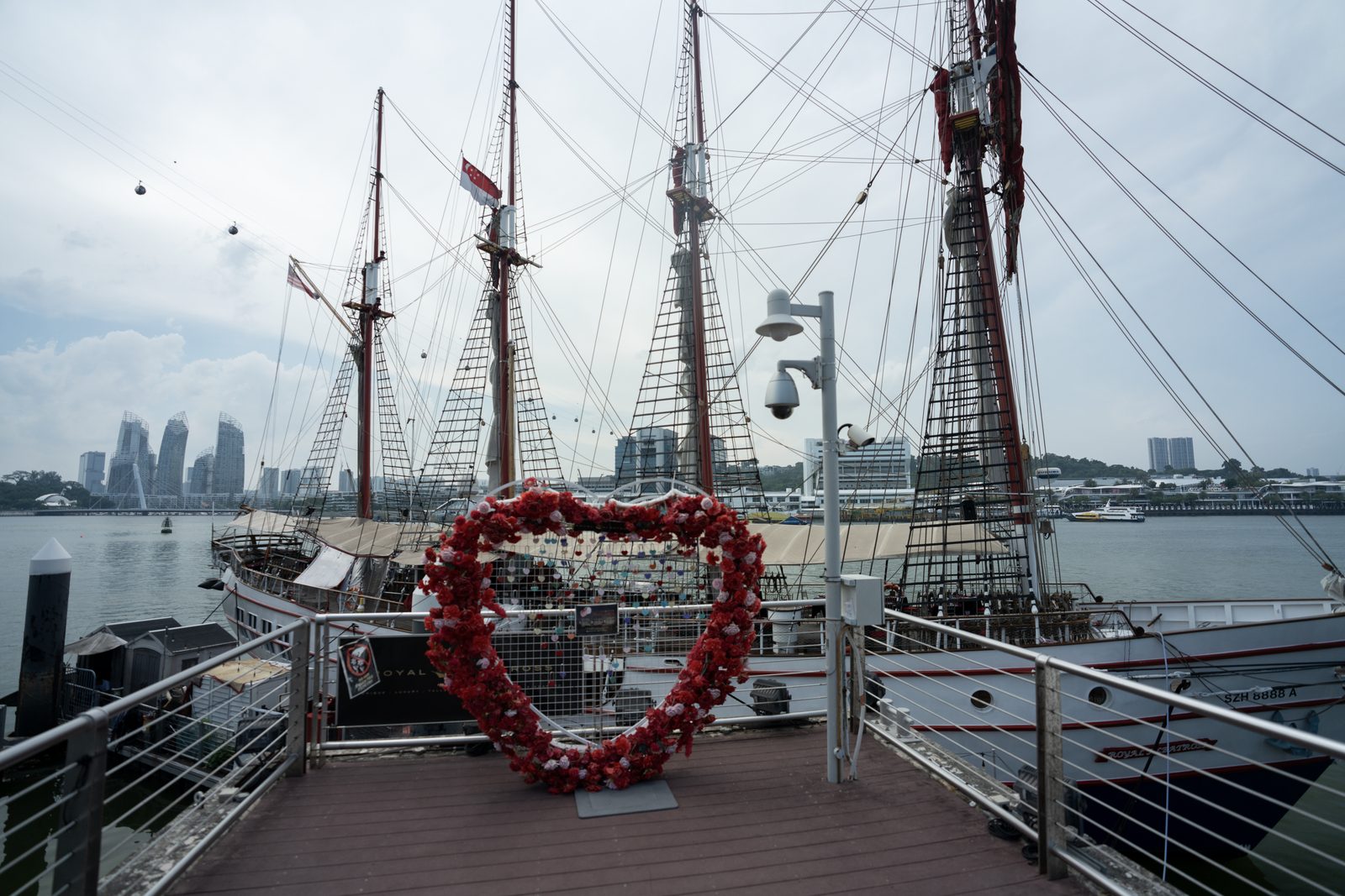 A heart-shaped frame of red flowers stands on a boardwalk before a moored tall ship, city towers hazy across the water.