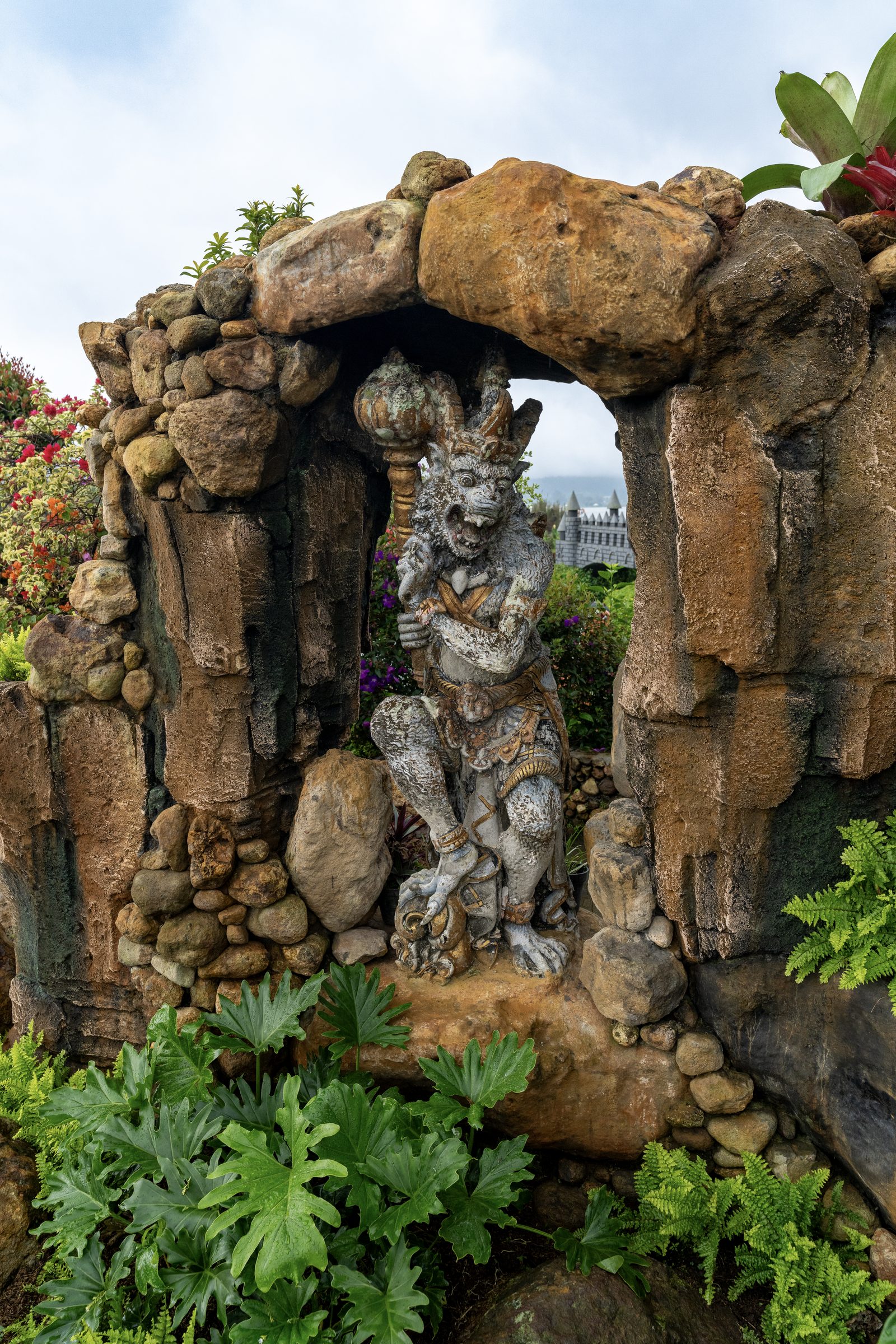 A carved stone figure stands under a rough rock arch in a garden, tropical leaves and ferns crowding the foreground.