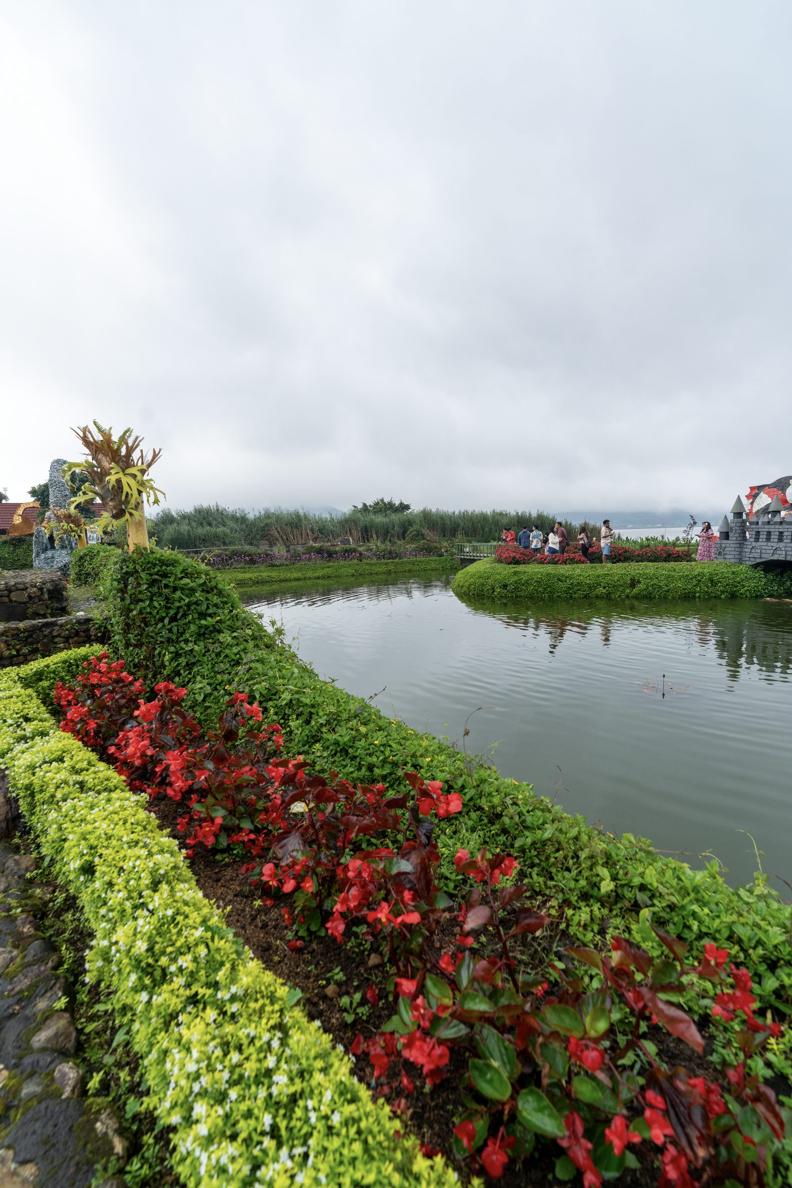 A long bed of red begonias and clipped hedge runs down to still lake water, low cloud sitting on the far shore.