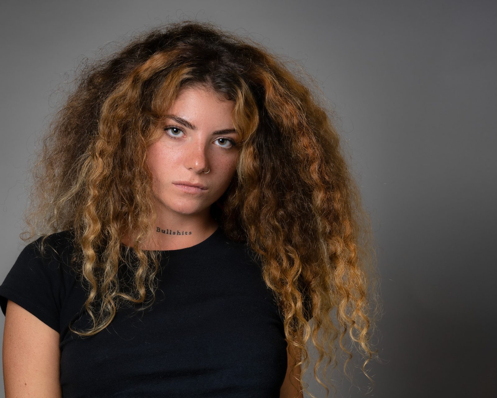 A woman with voluminous curly hair and a neck tattoo looks directly at the camera, wearing a black t-shirt against a grey studio backdrop