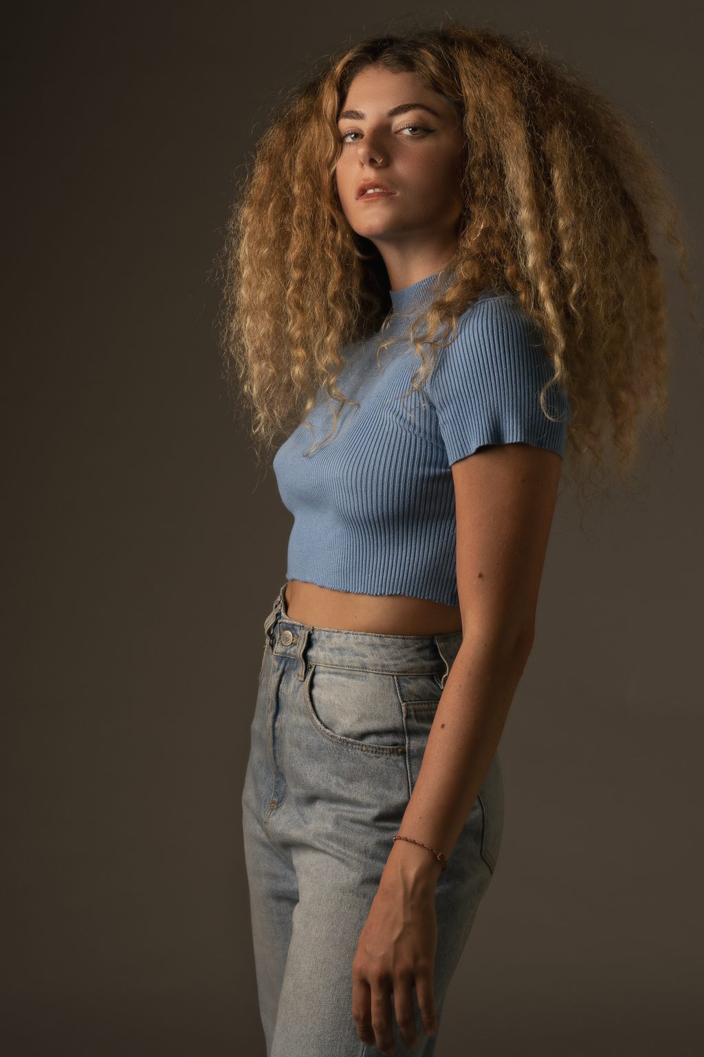 A woman with voluminous curly blonde hair stands in a pale blue ribbed crop top and light jeans against a warm brown studio backdrop