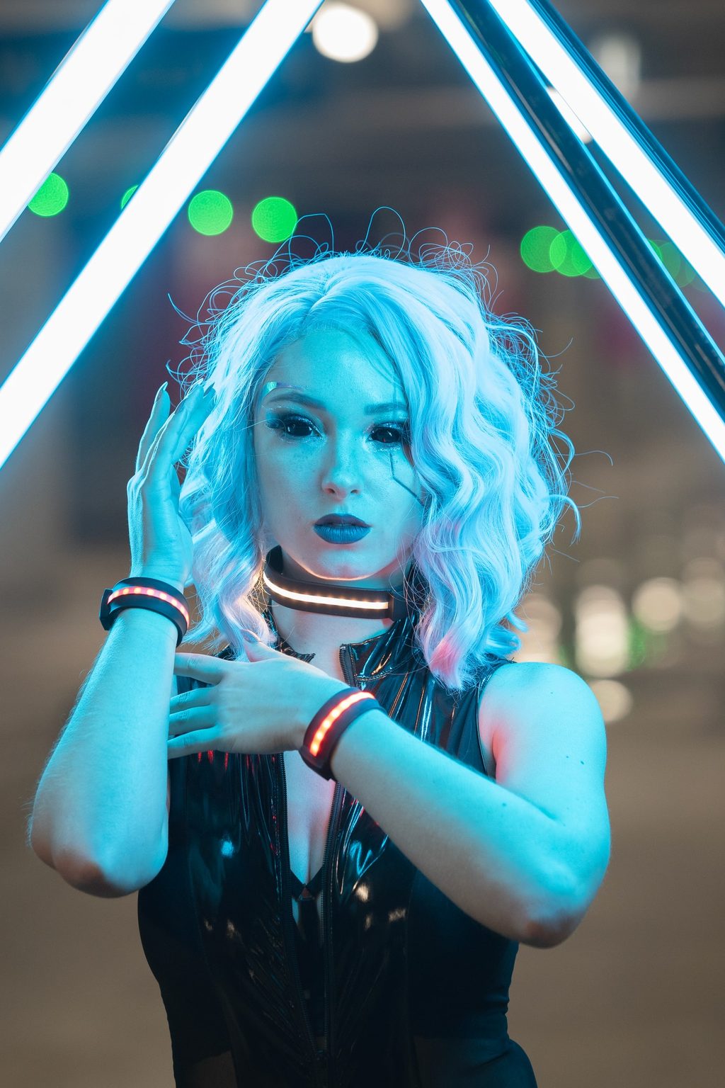 A cosplayer with pale wavy hair and a glossy black outfit stands under a triangular neon structure, LED accessories glowing red at wrist and throat