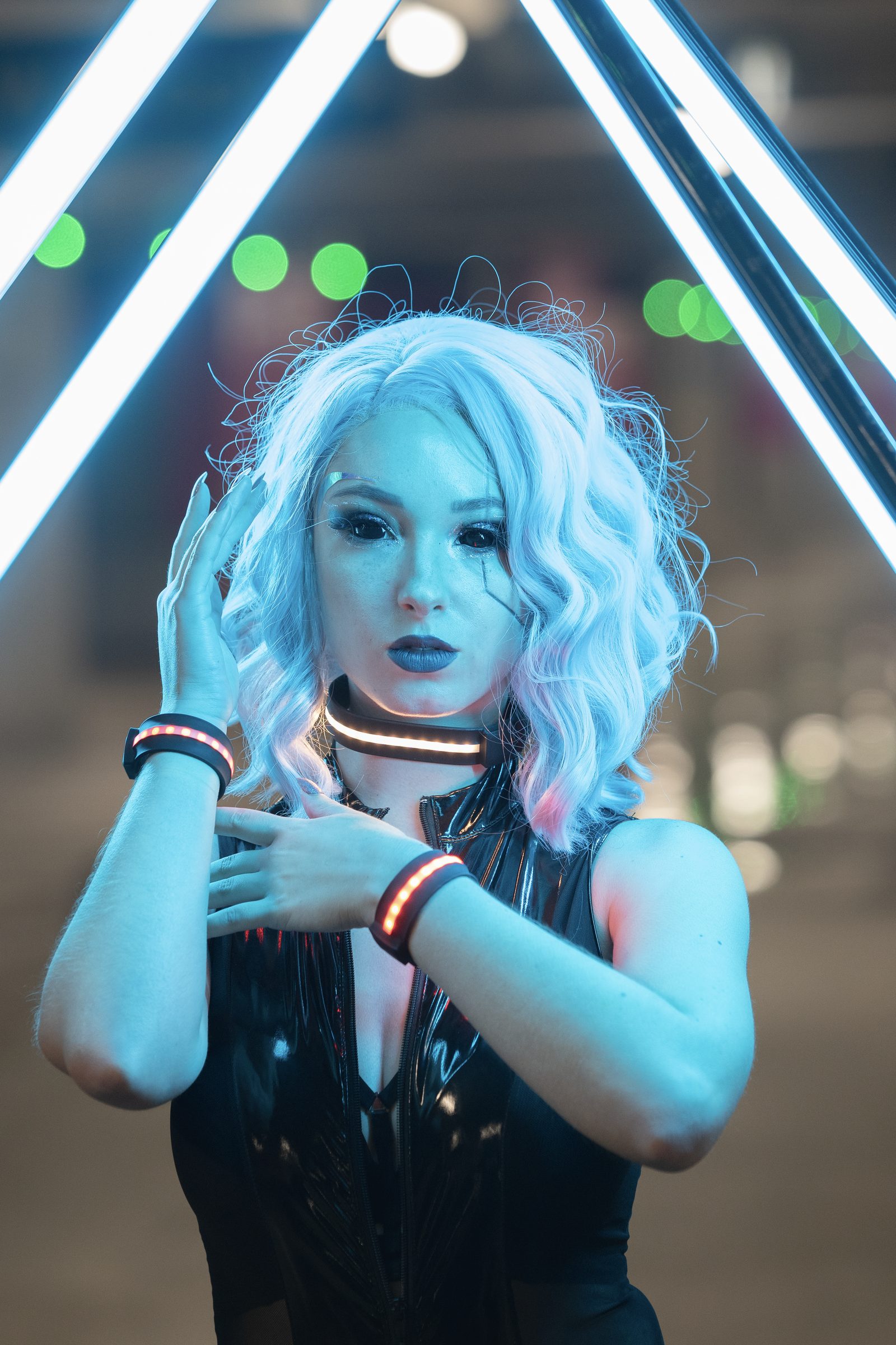 A cosplayer with pale wavy hair and a glossy black outfit stands under a triangular neon structure, LED accessories glowing red at wrist and throat
