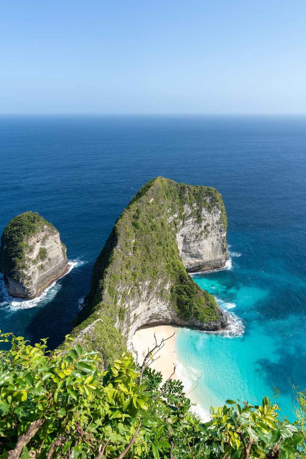 A forested limestone headland curves around a hidden white-sand cove, turquoise water breaking against the cliffs below.