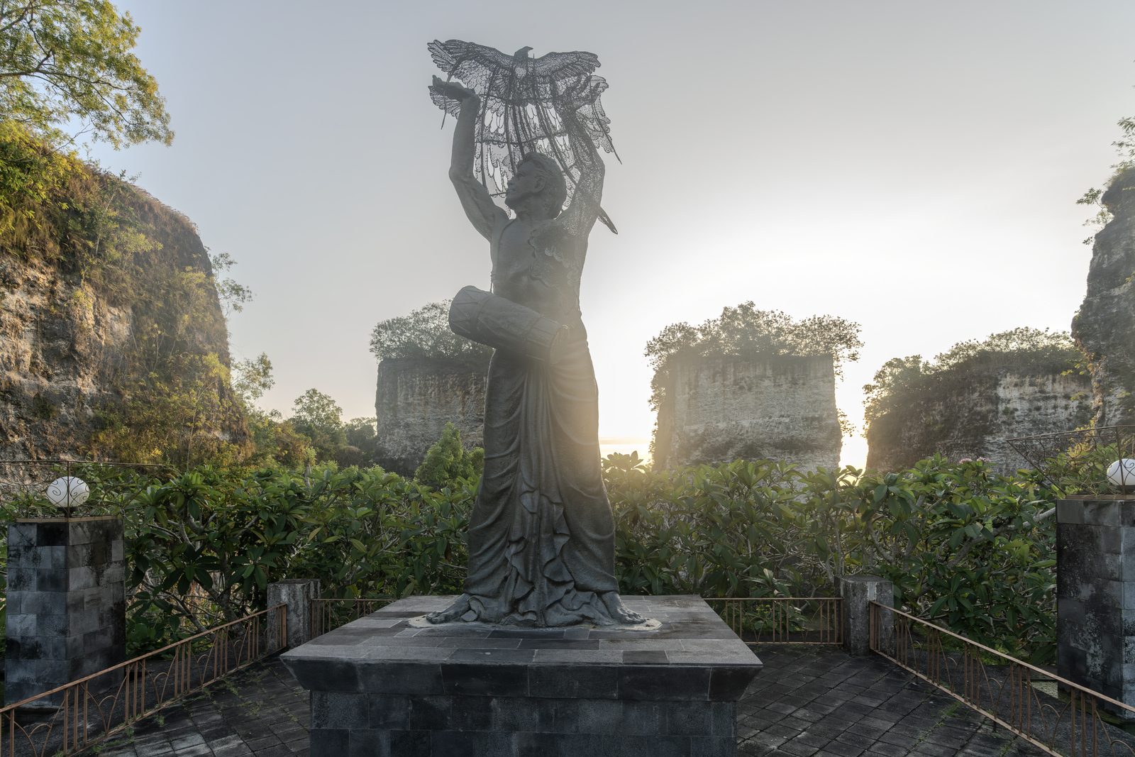 A stone figure raises carved wings above its head on a paved terrace, cut limestone walls and low sun behind.