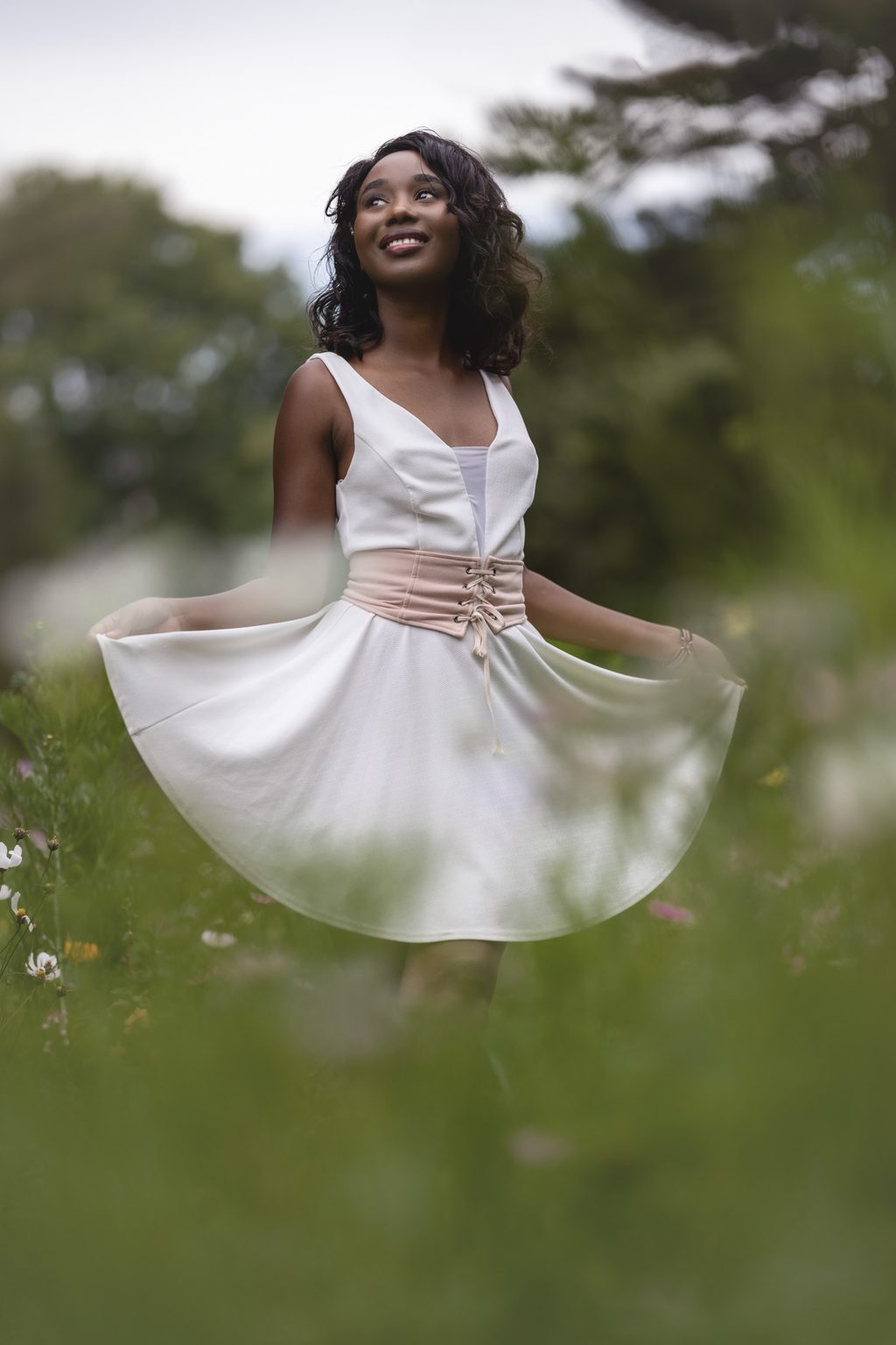 A woman in a white dress with a blush corset belt holds her skirt wide in a wildflower meadow, looking up and smiling