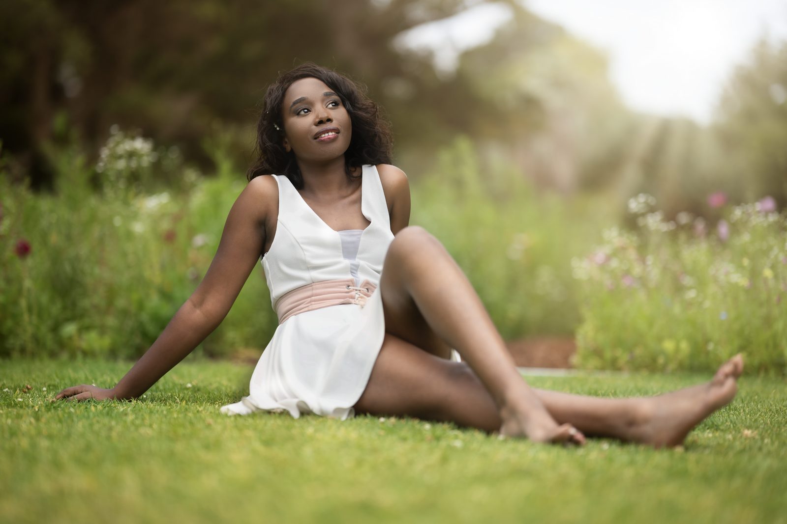 A woman in a white dress sits barefoot on mown grass with one knee raised, a flowering garden soft behind her