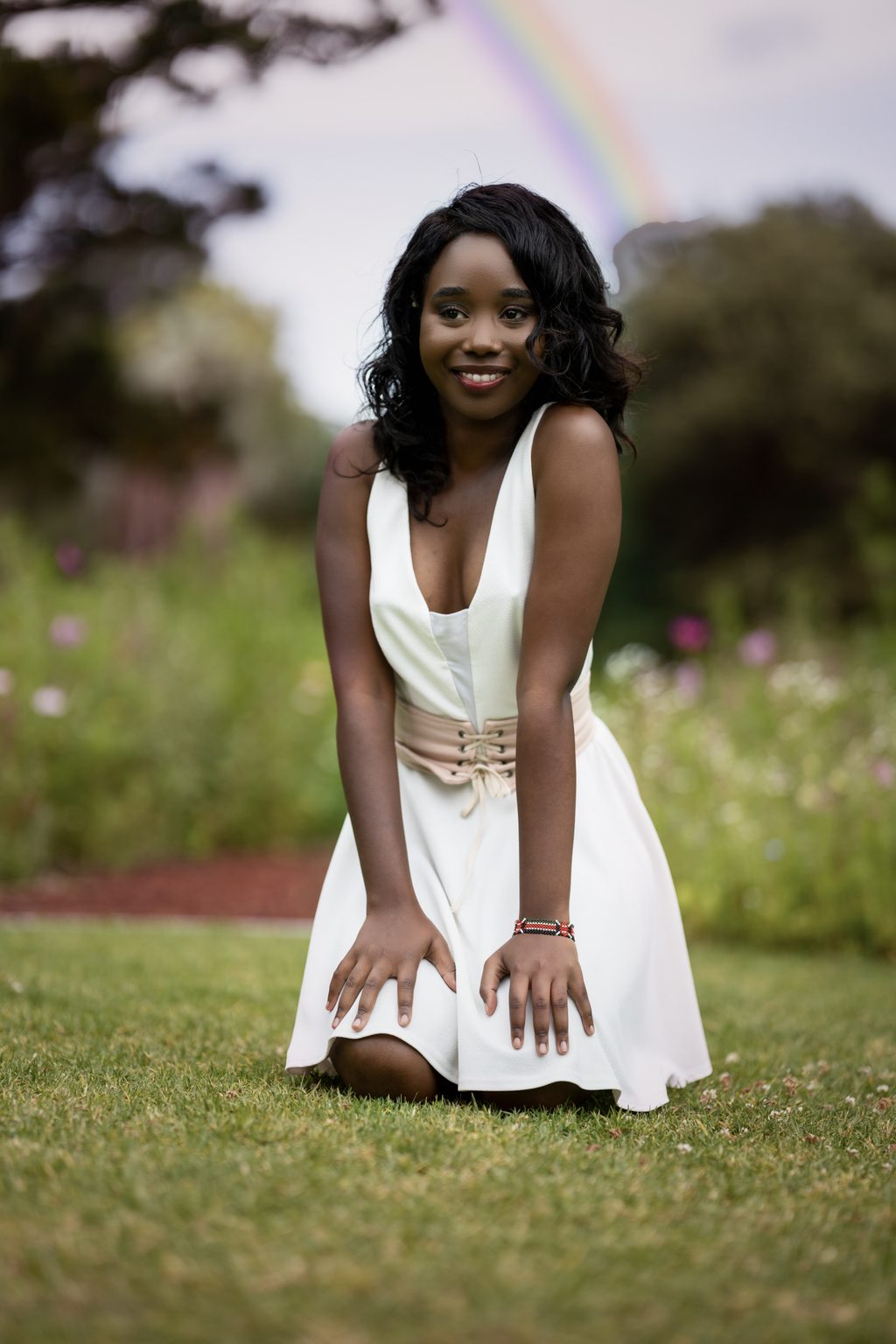 A woman in a white dress kneels on mown grass smiling to camera, a faint rainbow arcing over the trees behind