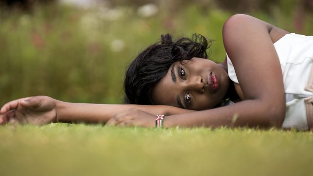 A woman lies on mown grass with her head resting on one outstretched arm, looking up into the camera