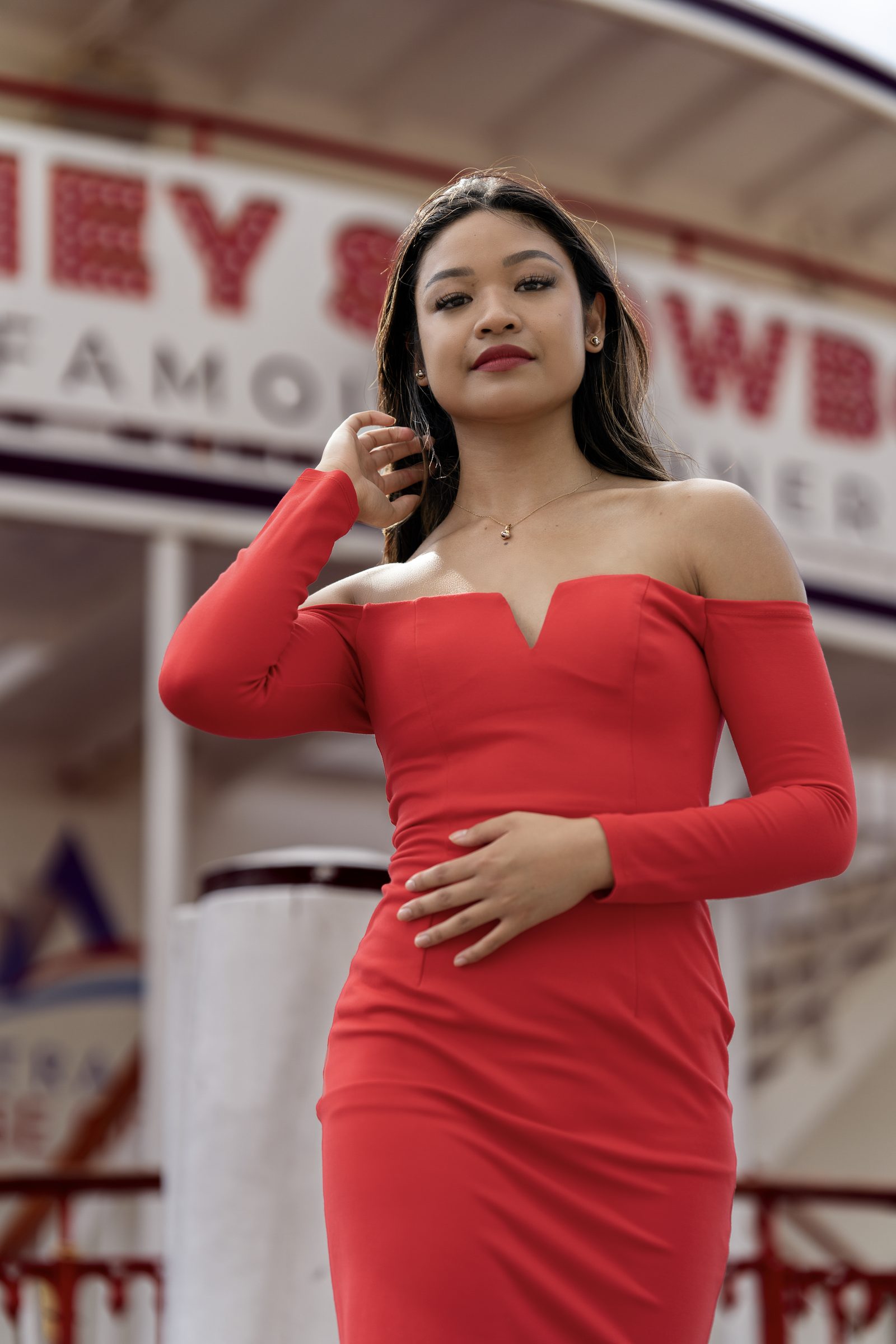 A woman in a red off-shoulder dress stands before a painted fairground sign, one hand lifted to her hair