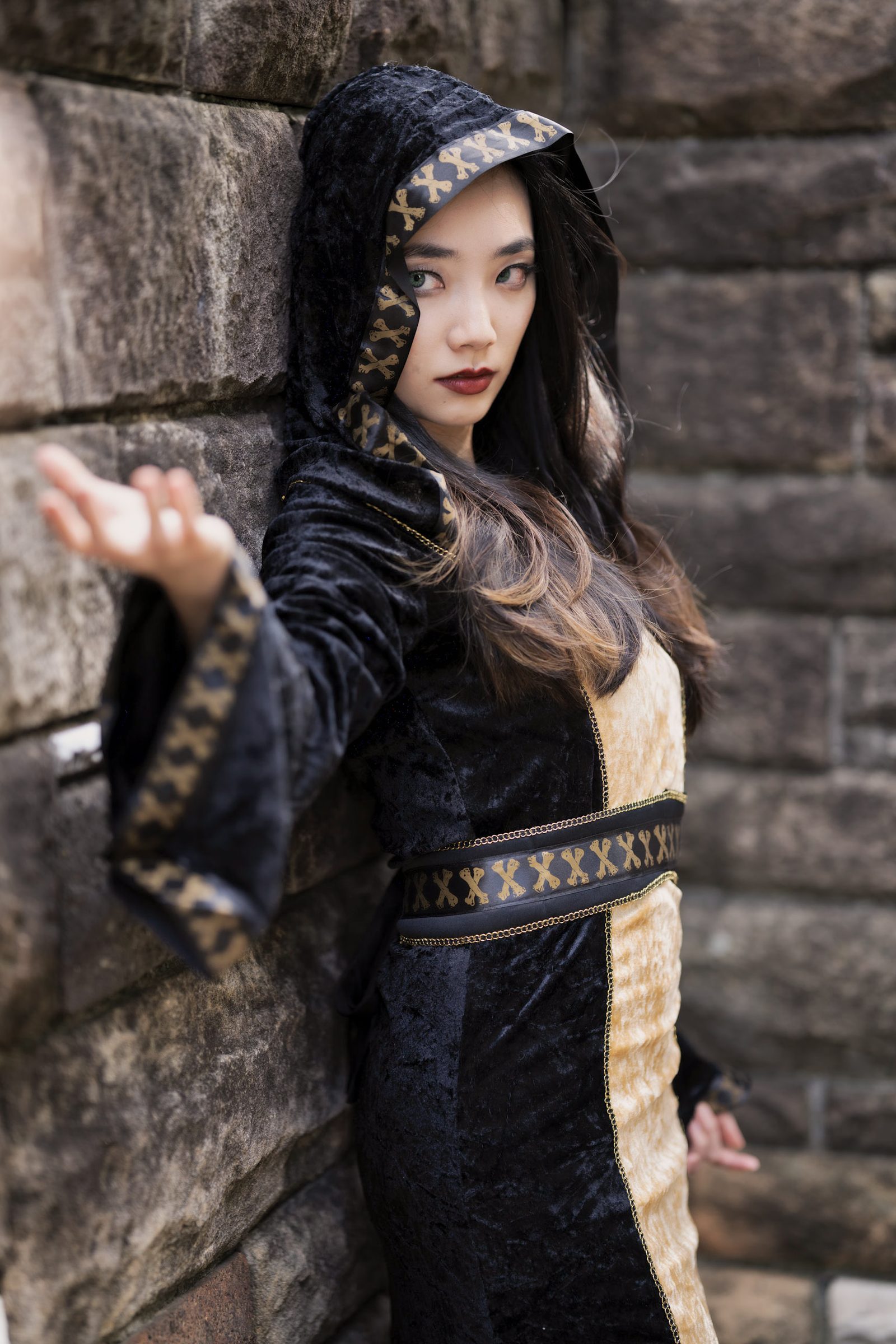 A woman in a hooded black and gold velvet costume with skull-and-crossbones trim reaches out one hand against a rough stone wall