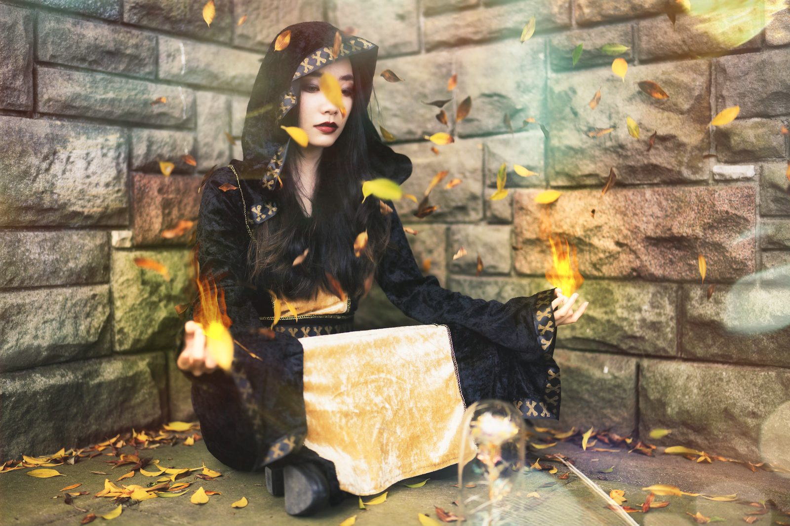 A figure in a dark hooded robe sits cross-legged against a stone wall holding flame in both hands, yellow leaves whirling around her