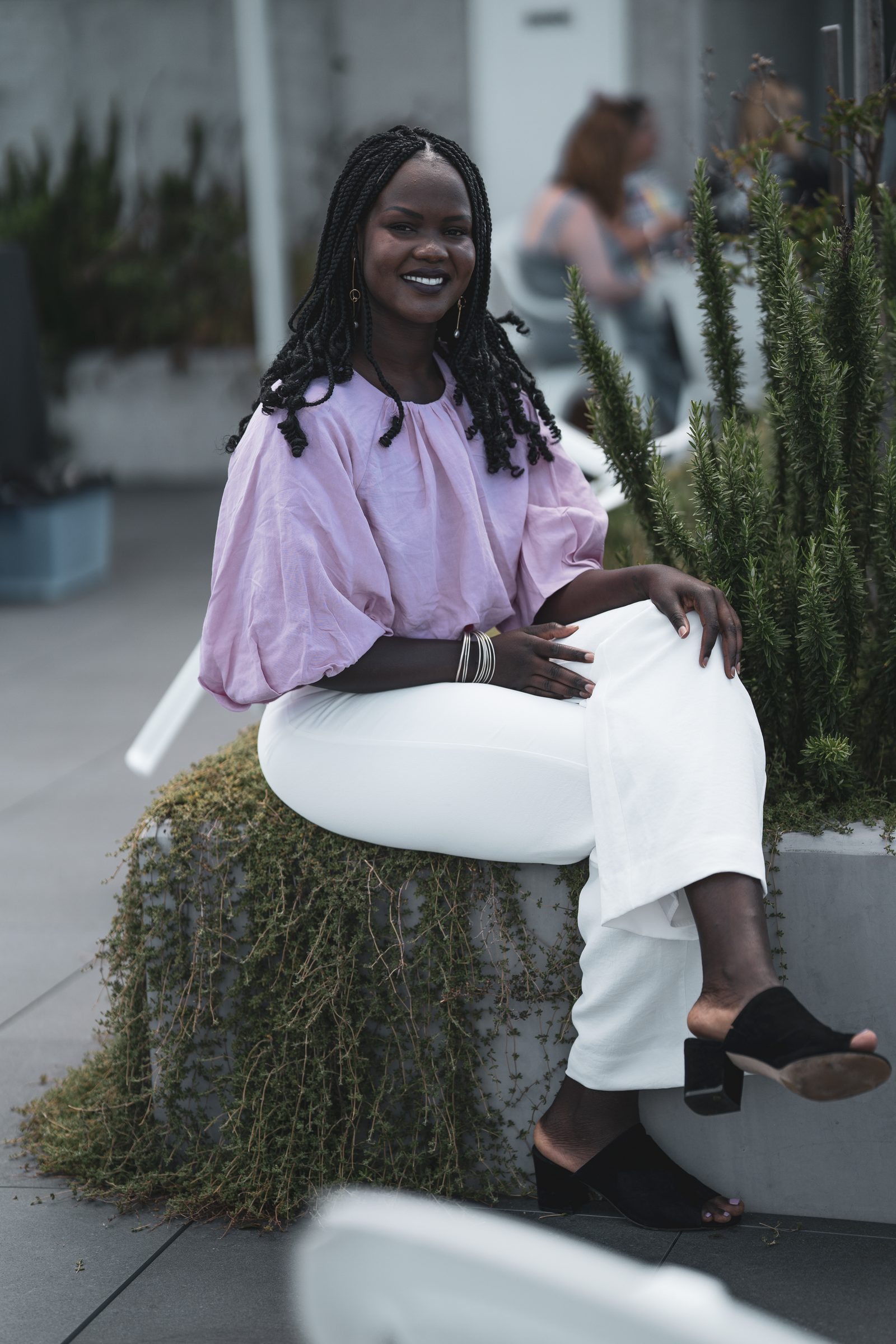 A woman with long braids in a lilac puff-sleeve blouse and white trousers sits on a planter edge, smiling to camera