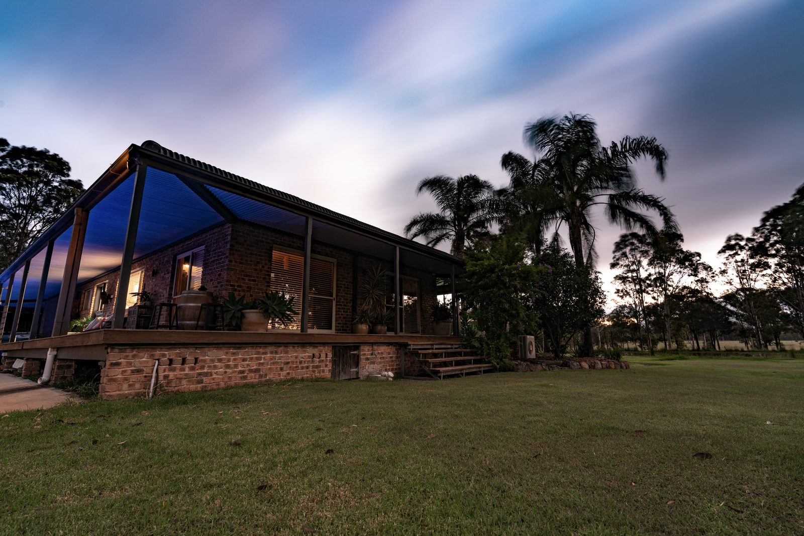 A low brick house with a deep verandah sits on mown grass at dusk, long-exposure cloud streaking above the palms.