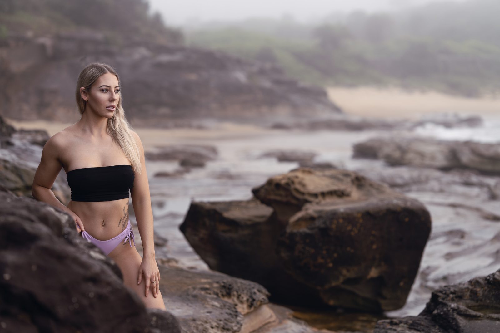 A woman in black and lilac swimwear stands among wet sandstone boulders, a misted headland and beach behind her