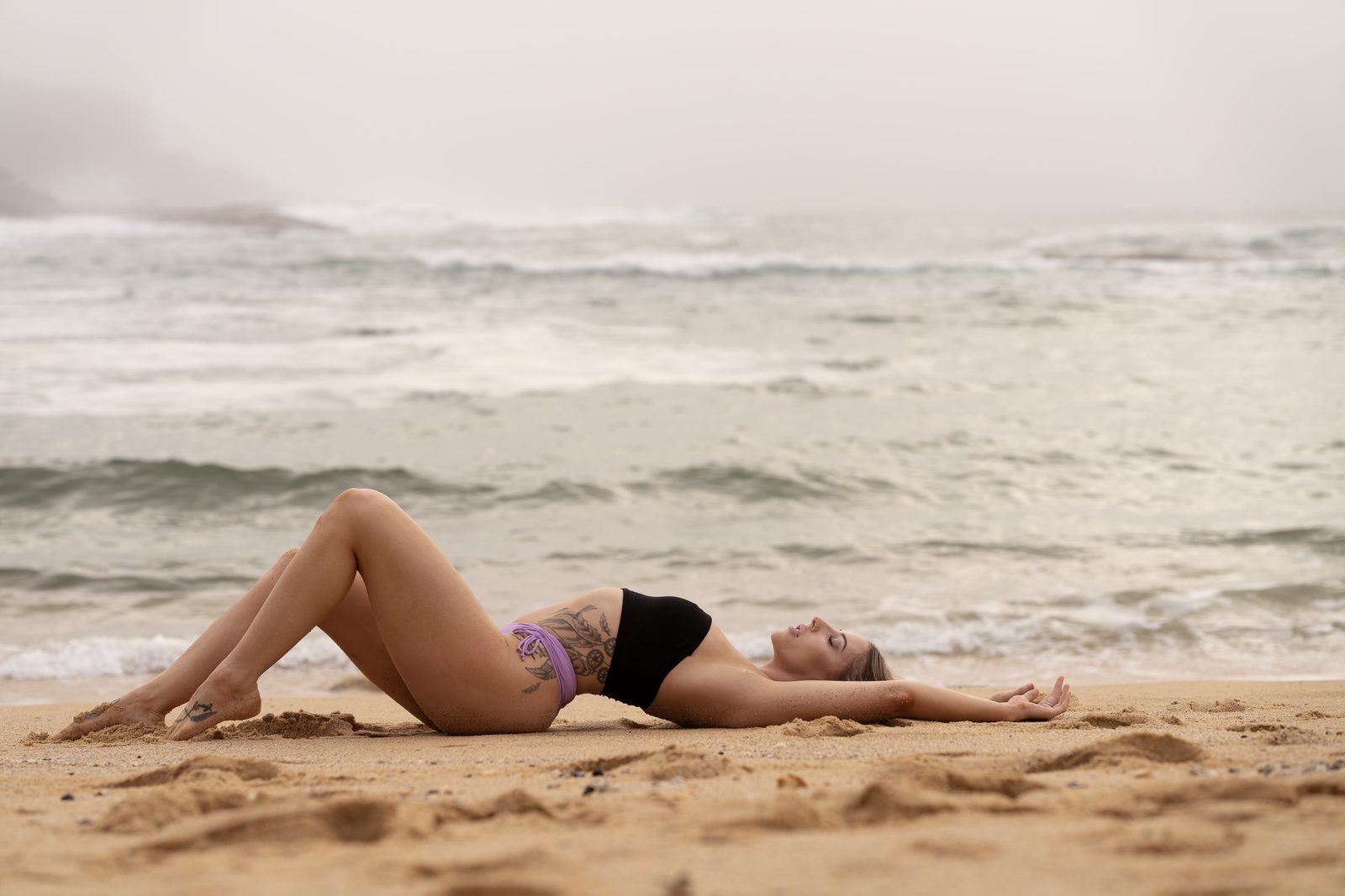 A woman in black and lilac swimwear lies back on the sand with her eyes closed, grey surf running behind her